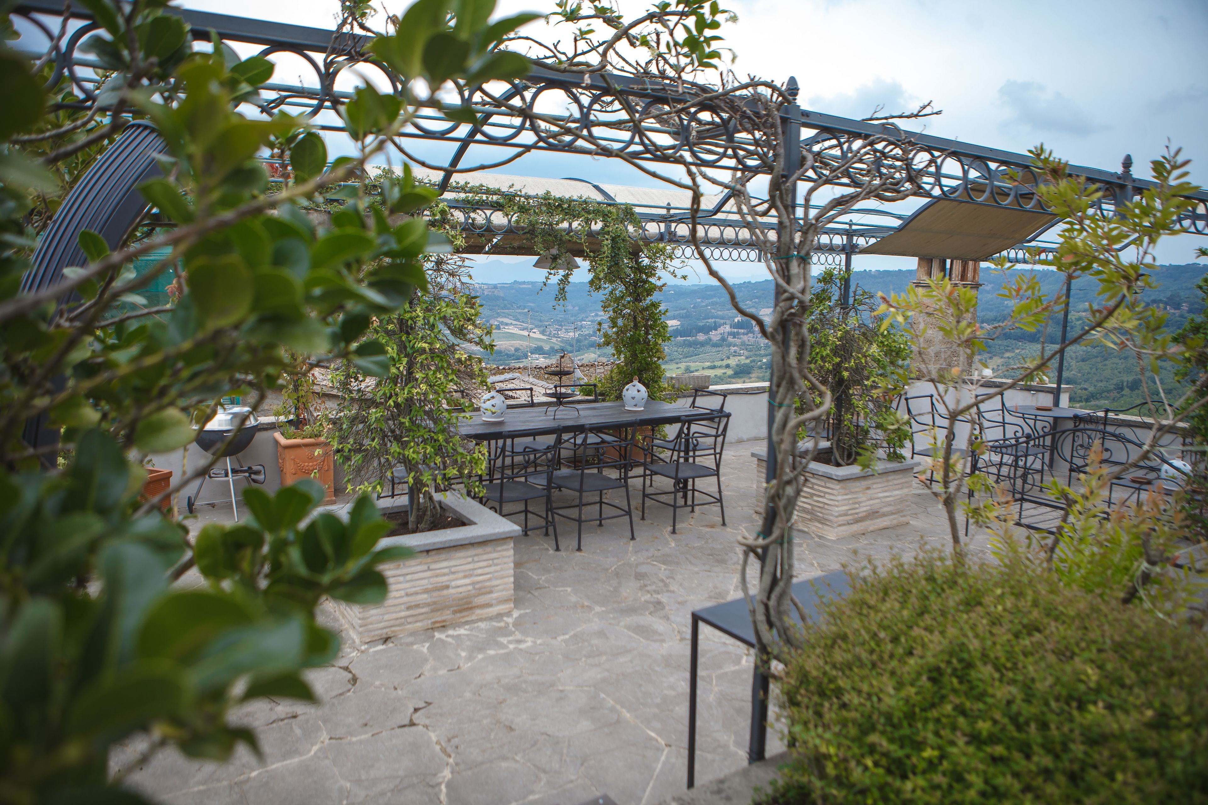 An outdoor patio with stone flooring, a table, and chairs, surrounded by greenery and overlooking a scenic landscape.