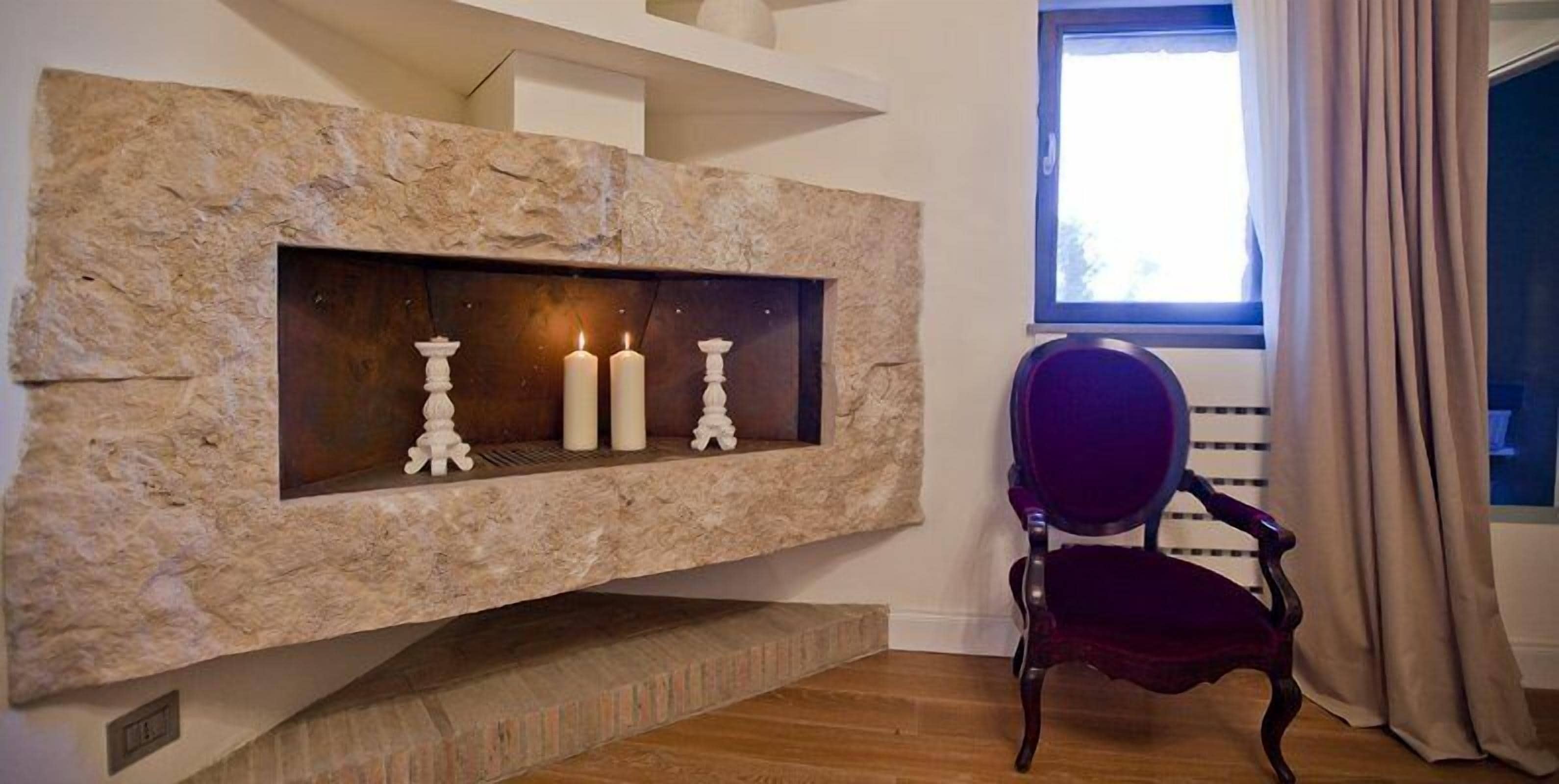 A stone fireplace with three candles, a dark wooden chair, and a window with curtains.