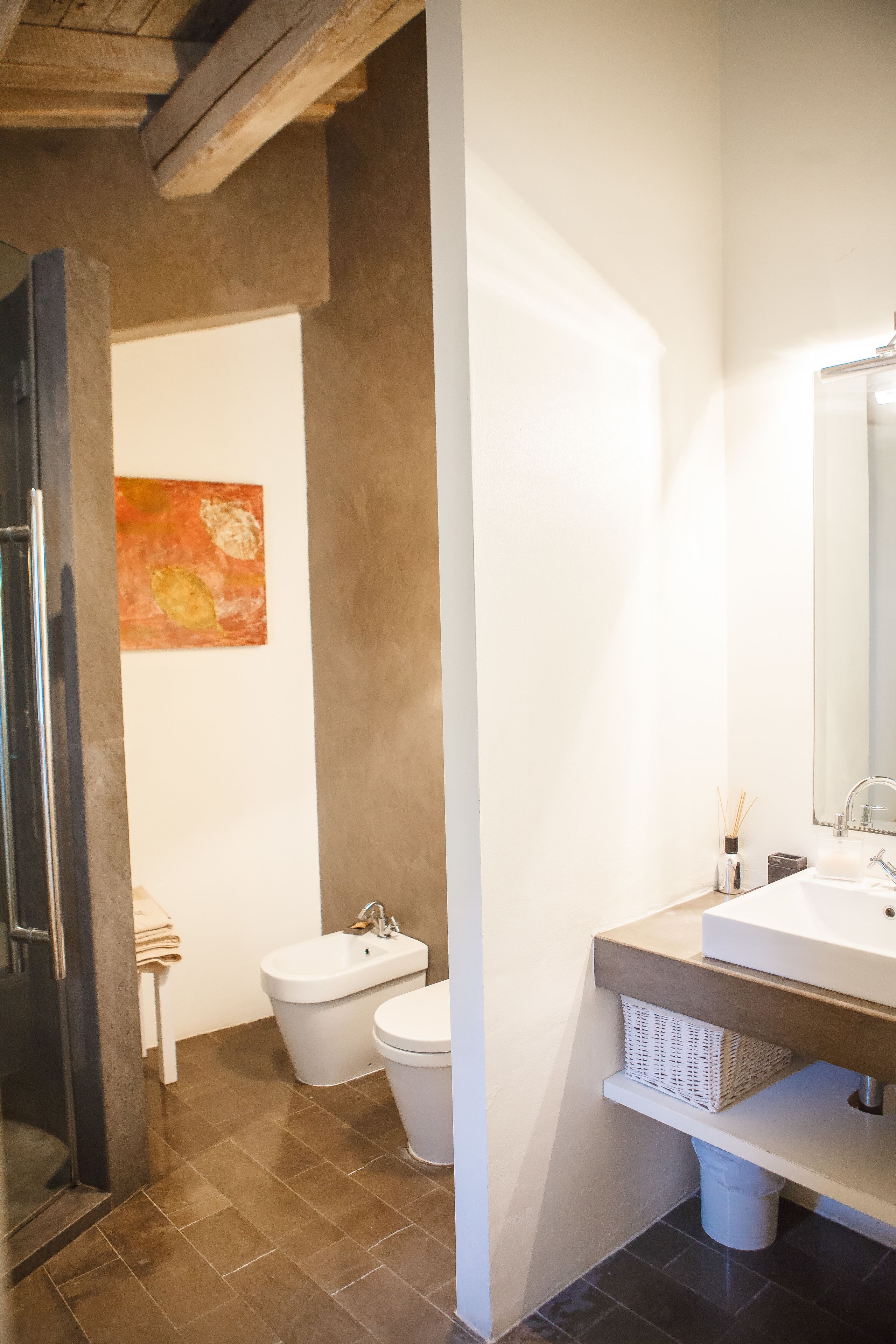 A bathroom with a shower, toilet, and sink. There is a painting on the wall and a basket under the sink.