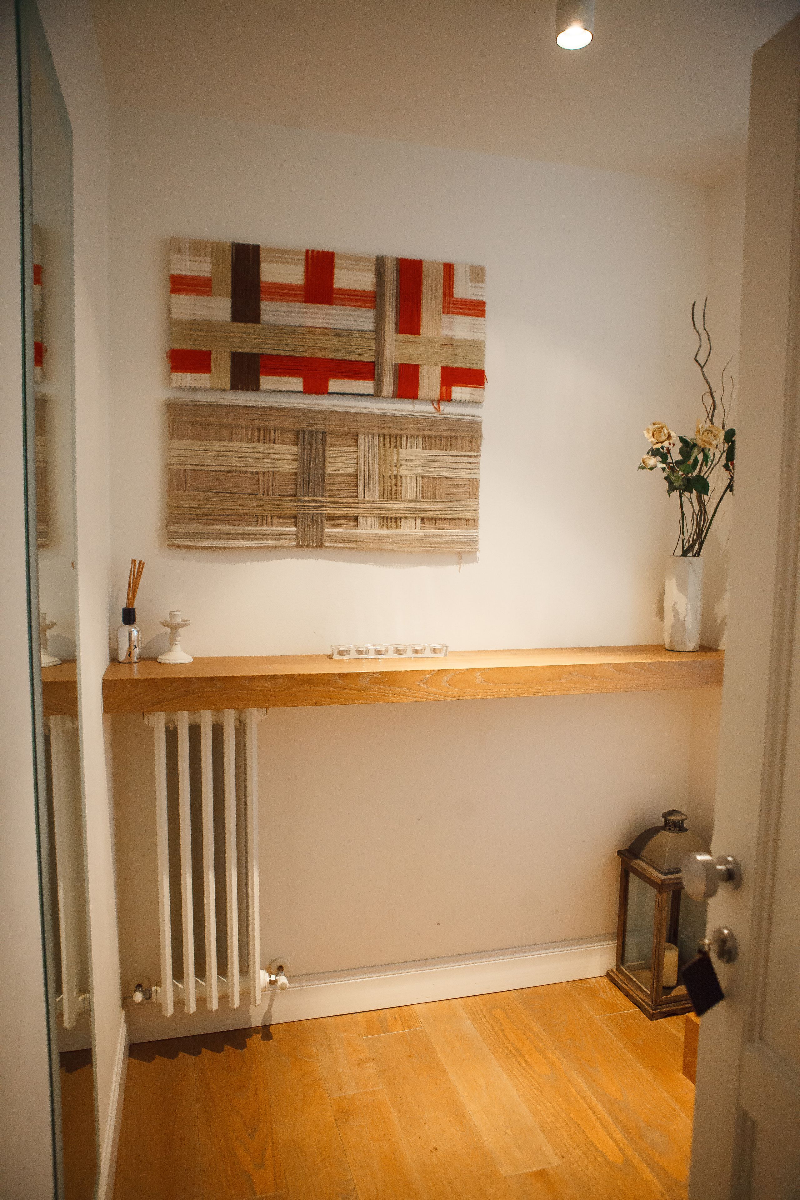 A hallway with a wooden floor, a radiator, a shelf with a decorative piece, and a wall-mounted artwork.