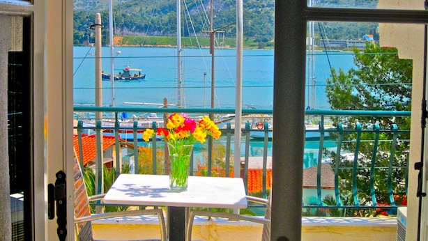 Un balcon avec une table et un vase de fleurs, donnant sur une marina où sont amarrés des bateaux, et sur un paysage montagneux.
