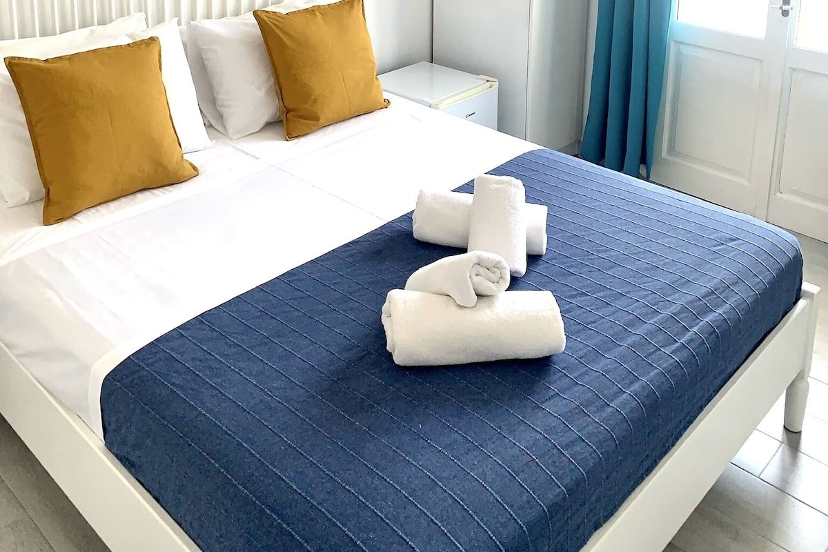 Quarto de hotel com uma cama, um quadro de uma paisagem costeira, um guarda-roupa e uma janela com cortinas.