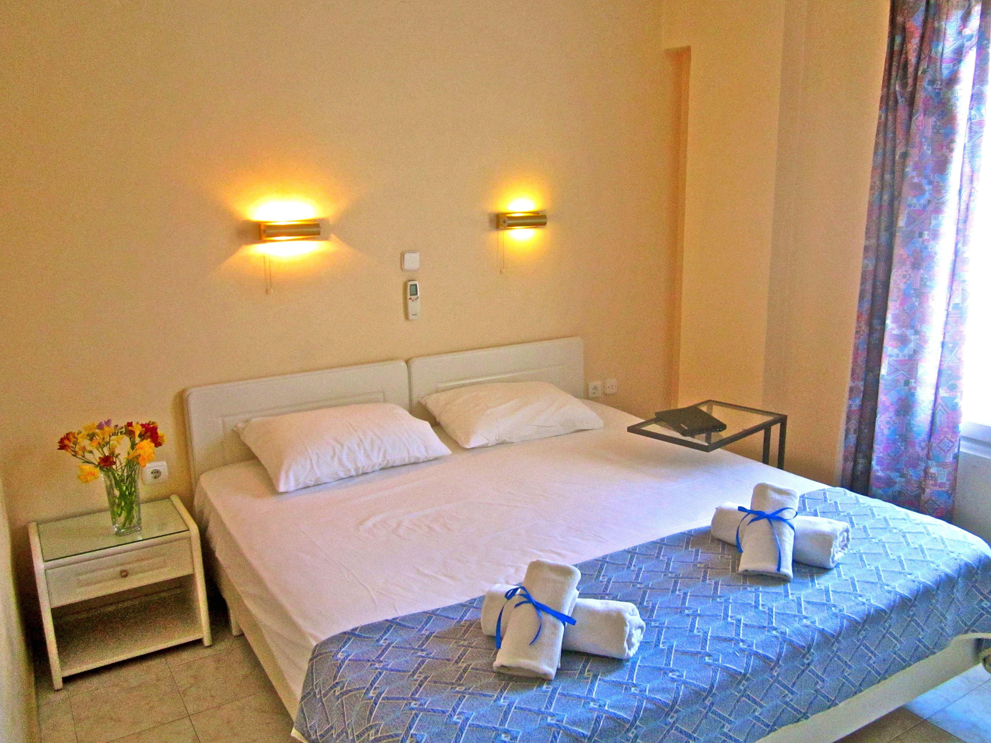 Ein Hotelzimmer mit einem Bett, zwei Nachttischlampen, einem Nachttisch mit Blumenvase und einem Fenster mit Vorhängen.