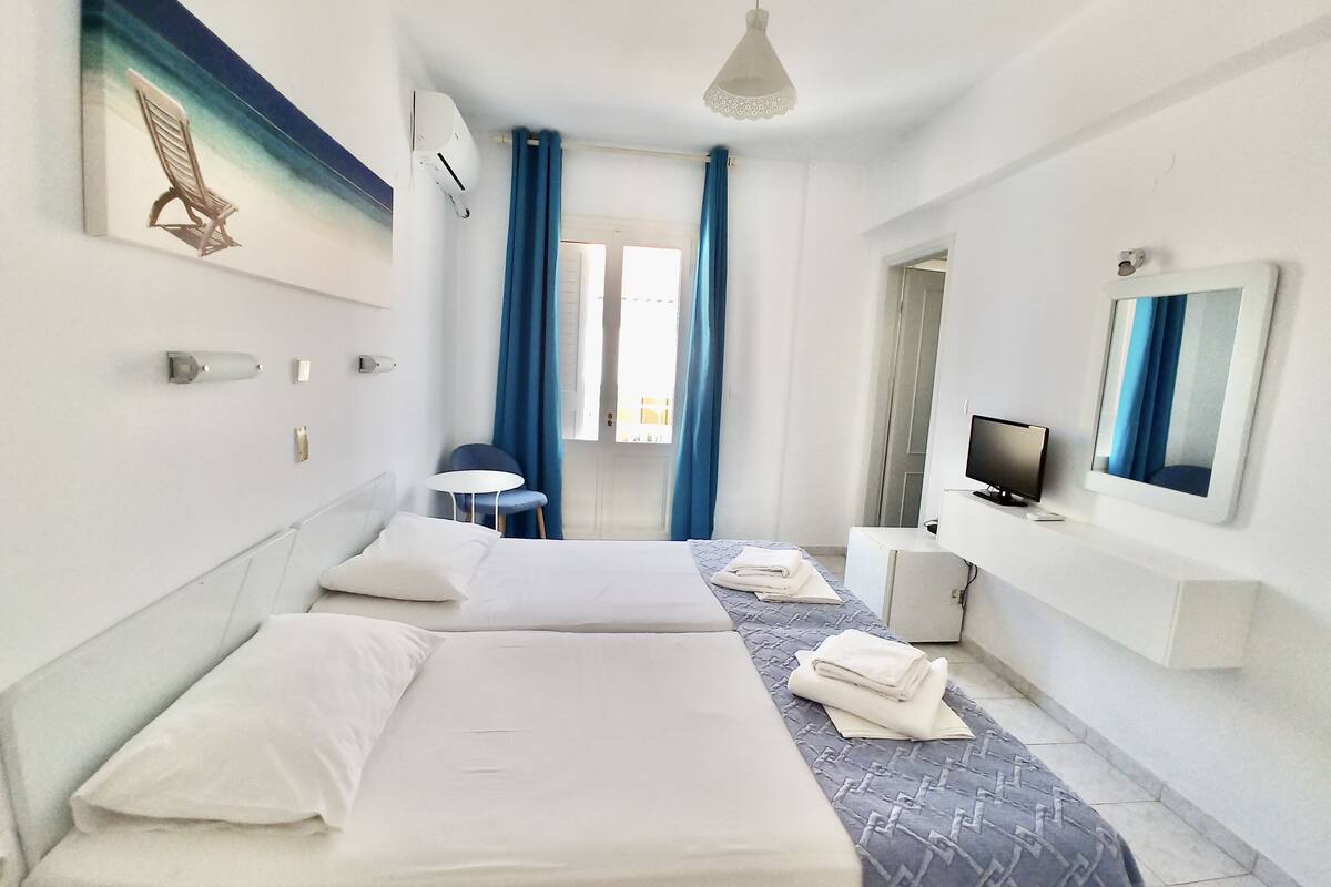 Quarto de hotel com duas camas, uma mesinha, uma cadeira, uma televisão e um espelho.