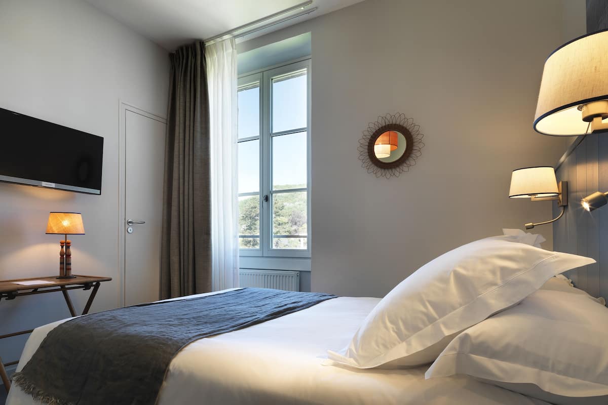 Un lit bien fait, avec des oreillers blancs et un plaid gris, une lampe de chevet, un miroir fixé au mur et une fenêtre avec des rideaux.