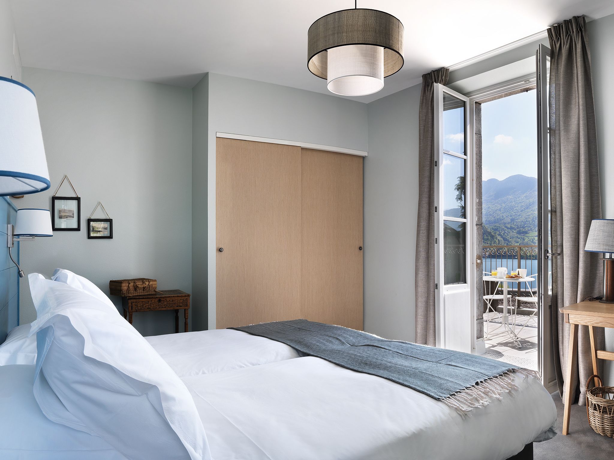 A bedroom with a bed, a hanging light fixture, a wardrobe, a small table with a chair, and a view of the outdoors.