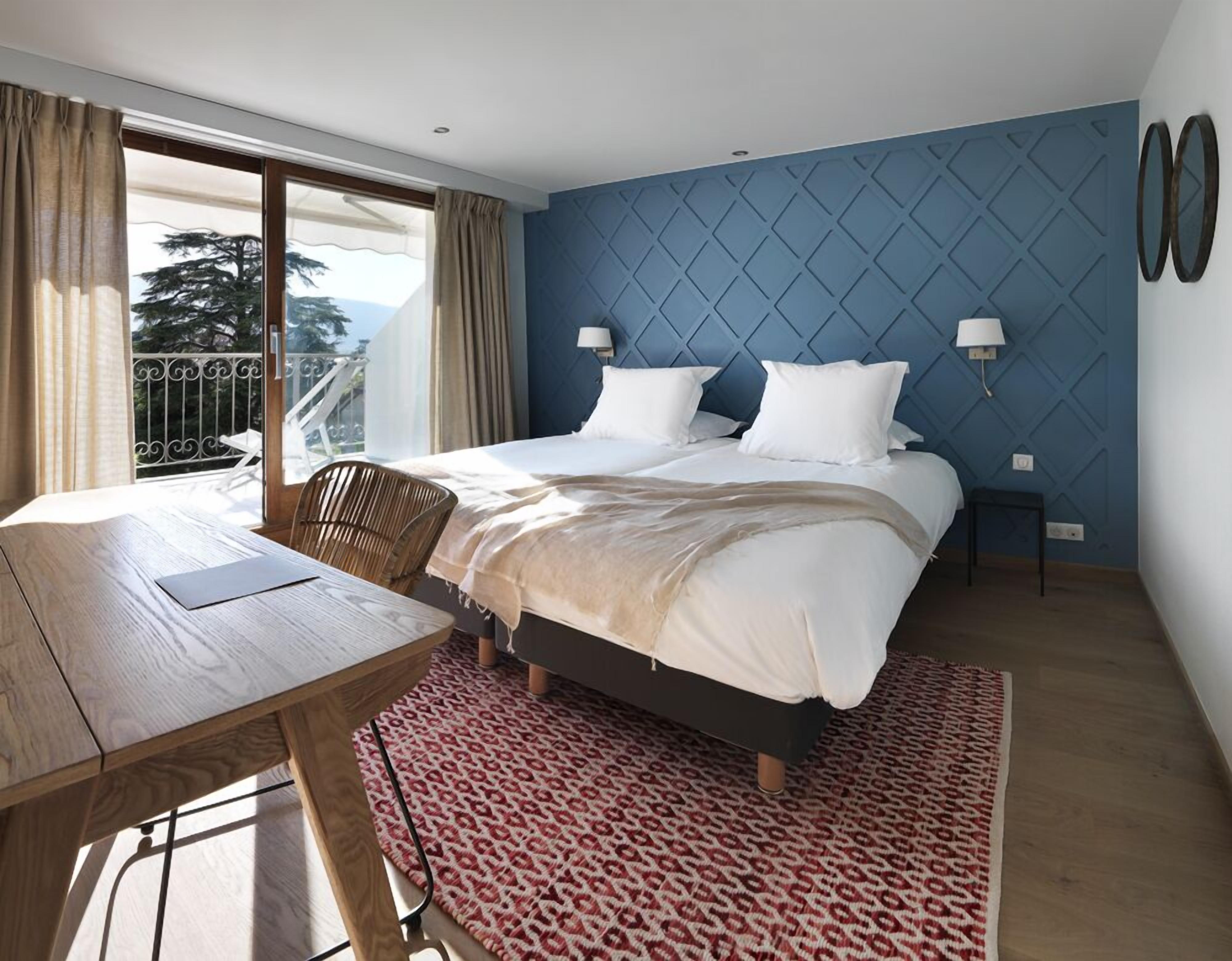 A hotel room with a bed, a desk, and a chair. There is a balcony with a railing and a view of trees.