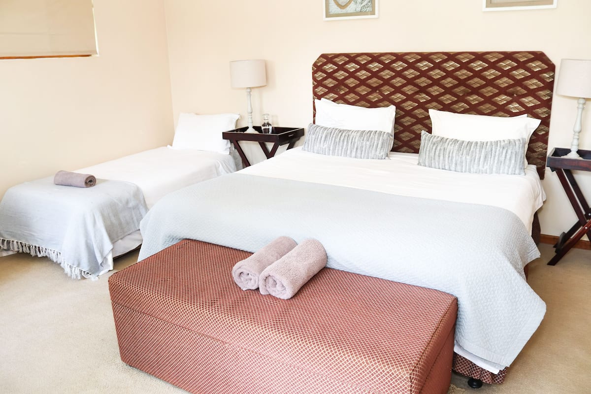 A hotel room with a large bed, two smaller beds, a red woven bench, and a small table with a lamp.