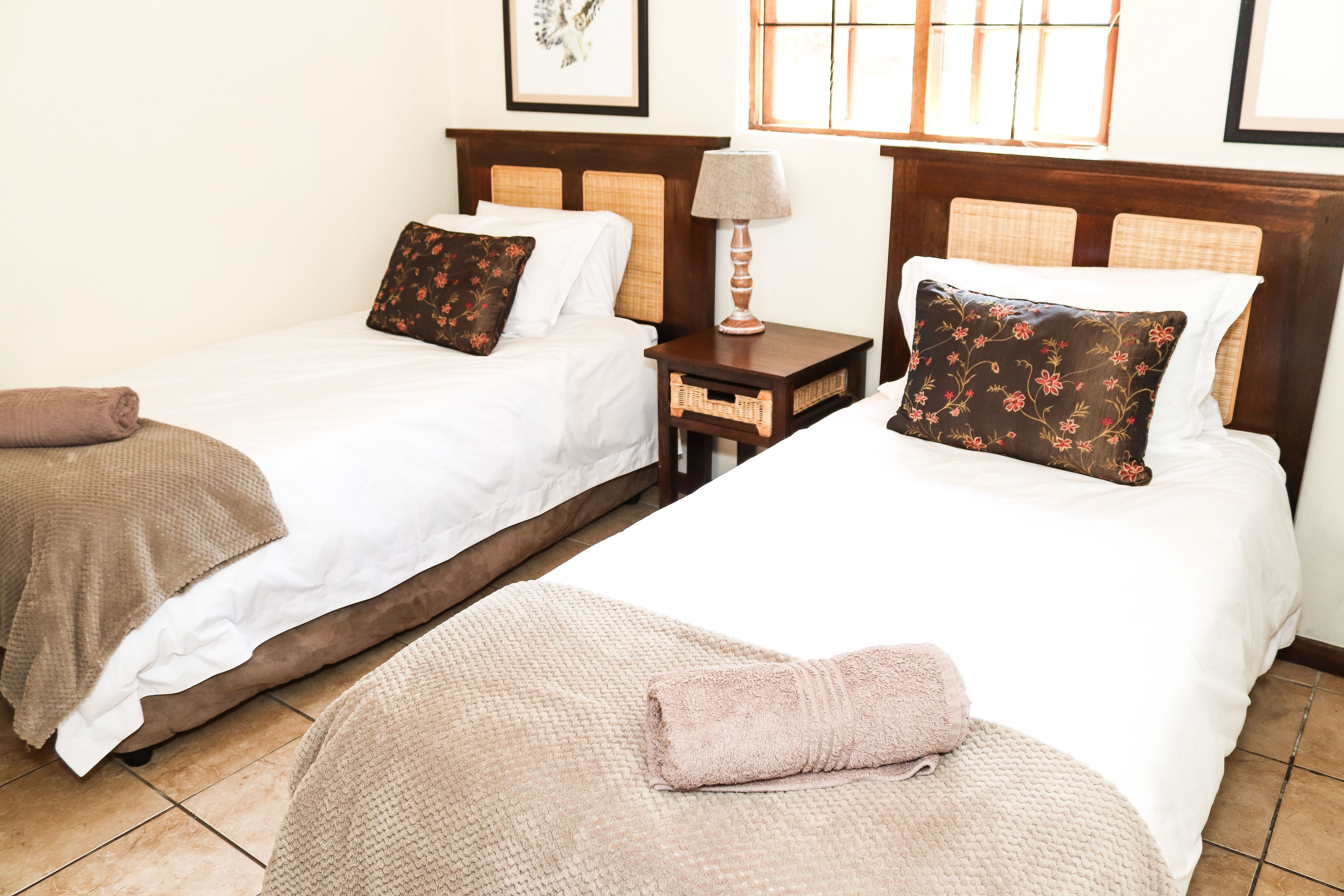 A hotel room with two beds, each with a pillow and a blanket. There is a nightstand with a lamp between the beds.