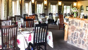 A restaurant with wooden chairs and tables, stone bar counter, and large windows with curtains. - The Old Hatchery (Drakensberg Gardens)
