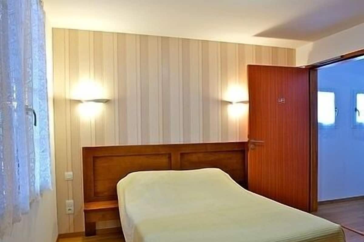 A room with a wooden bed, two wall-mounted lights, a wooden door, and a view through an open door.