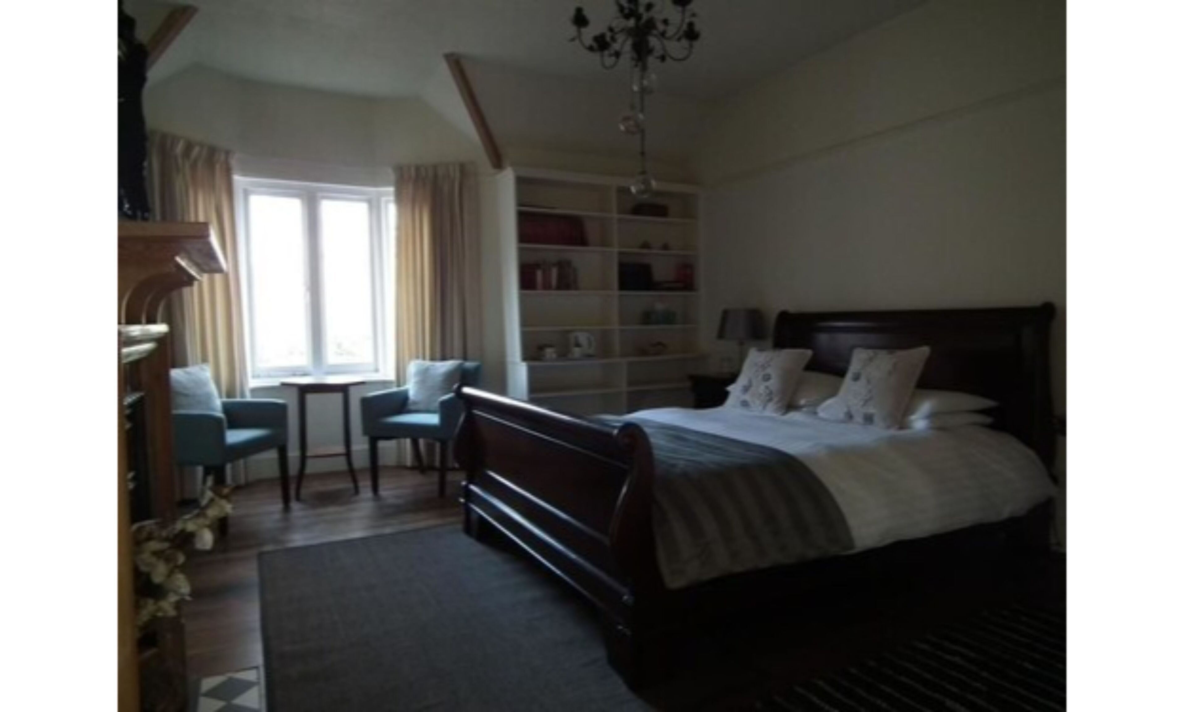A bedroom with a large bed, a desk, and a chair. There is a bookshelf and a chandelier.
