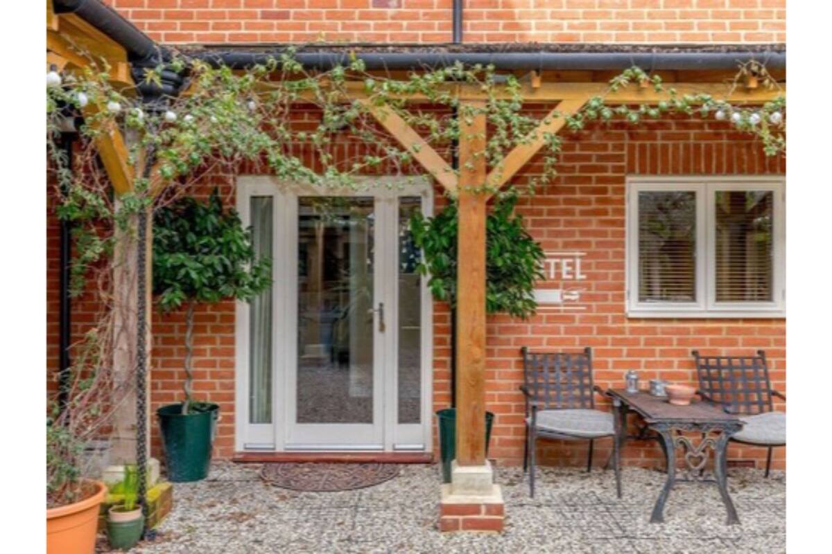 A brick building with a glass door entrance, a wooden pergola with climbing plants, and a small outdoor seating area with a table and chairs.