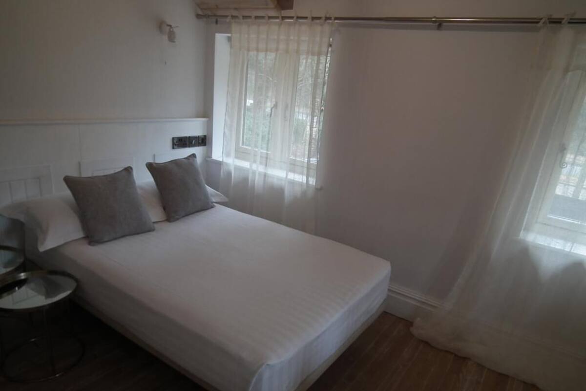 A neatly made bed with white linens and two pillows, a bedside table with a lamp, and a window with sheer curtains.