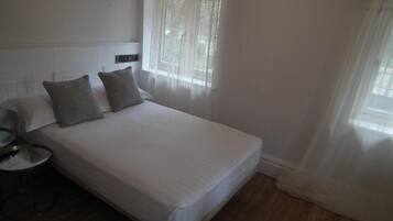 A neatly made bed with white linens and two pillows, a bedside table with a lamp, and a window with sheer curtains.
