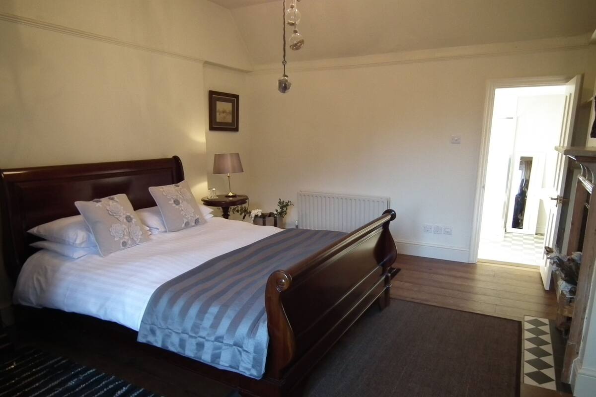 A bedroom with a large bed, a nightstand with a lamp, a radiator, and a view of the hallway.