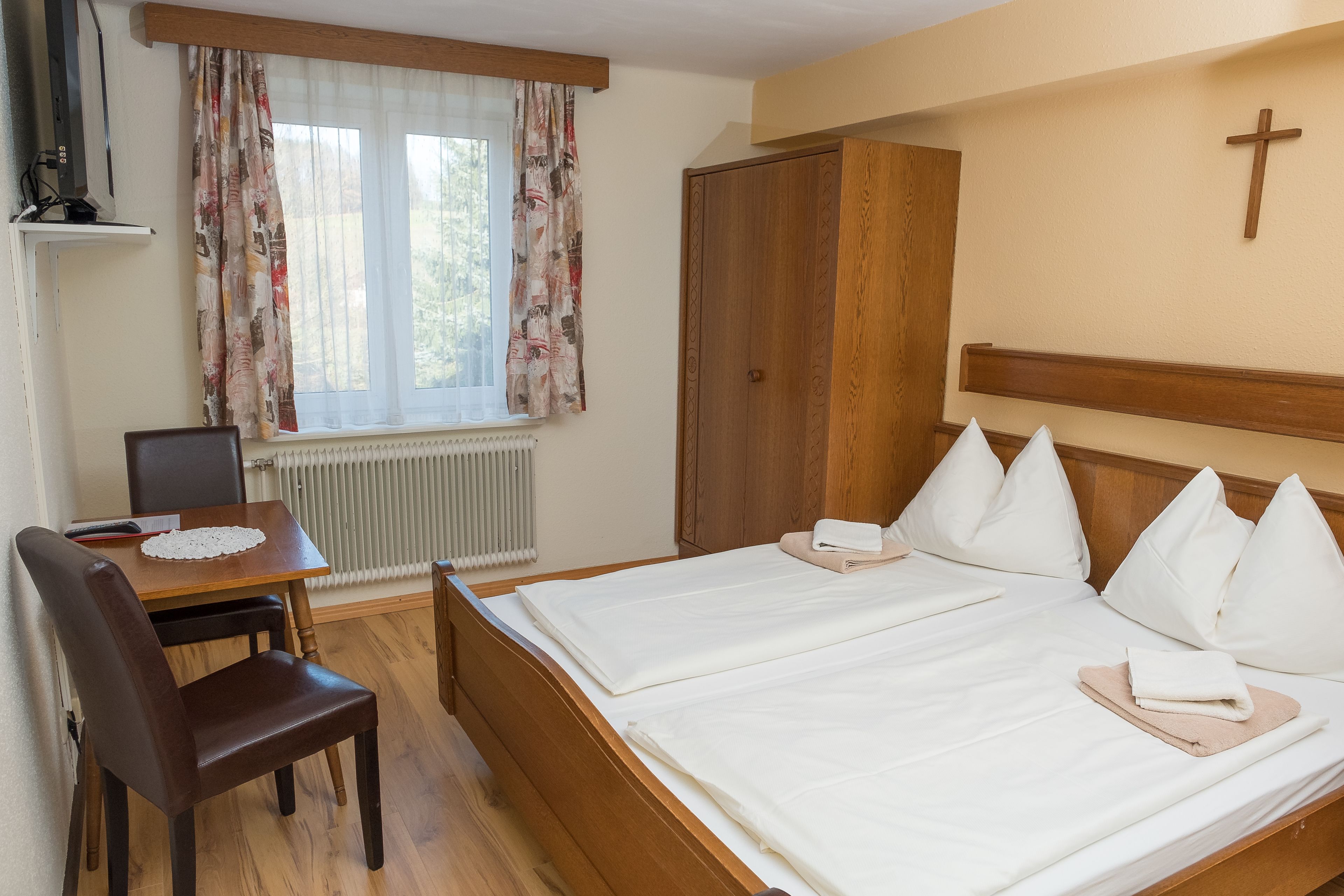 A double bed with white linens, a wooden headboard, and a cross on the wall. There is a wooden desk with a chair, a radiator, and a window with curtains.