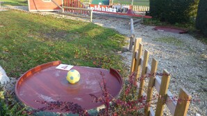 Un parque infantil con una pelota roja sobre una pista circular, una valla de madera y un campo verde al fondo.