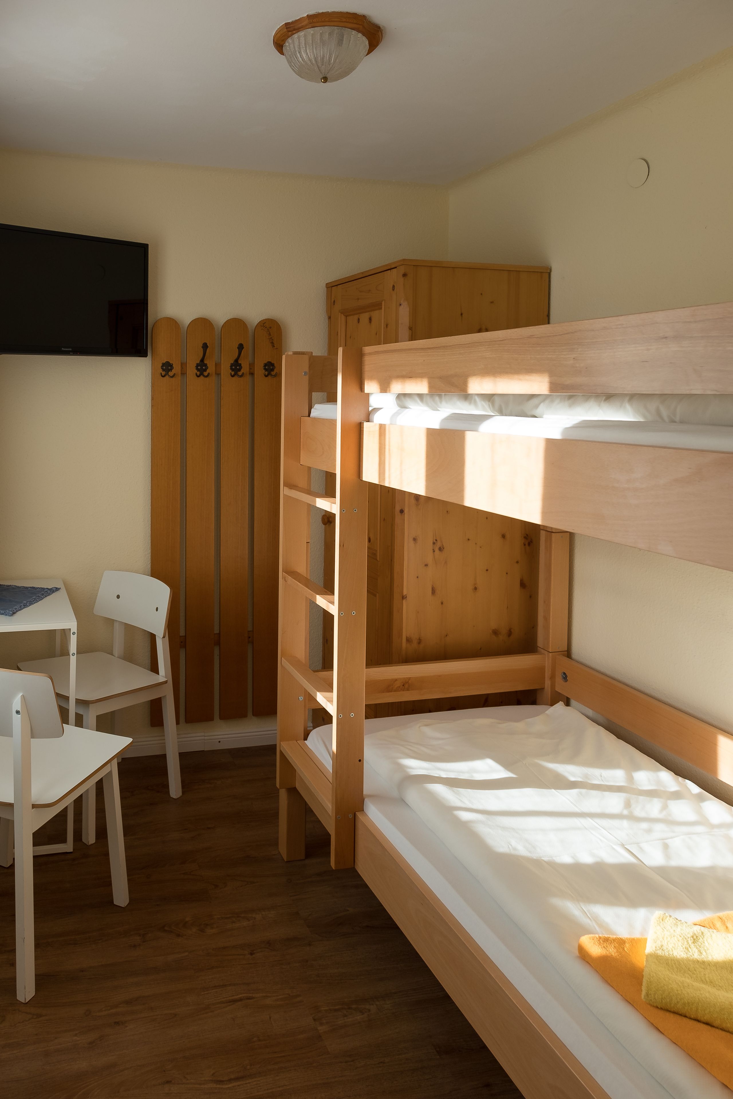 A bunk bed room with a desk and chairs, wooden wardrobe, and a television on the wall.