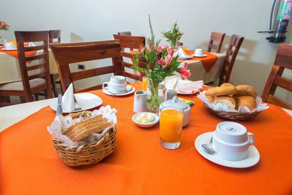A breakfast setting with a table set for two, including bread, pastries, a glass of orange juice, and a vase of flowers.