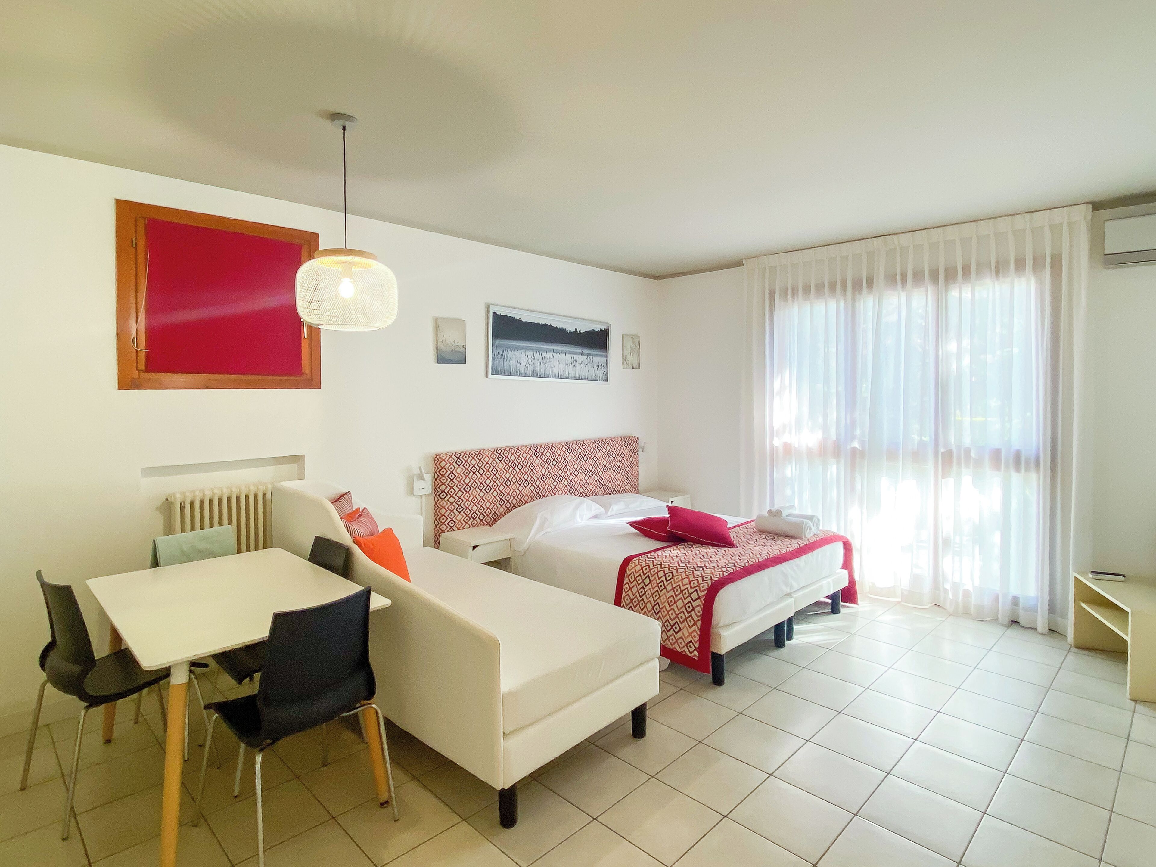 Camera d'albergo con un letto, una scrivania e una sedia. Si vede una finestra con tende, un termosifone e un condizionatore d'aria a parete.