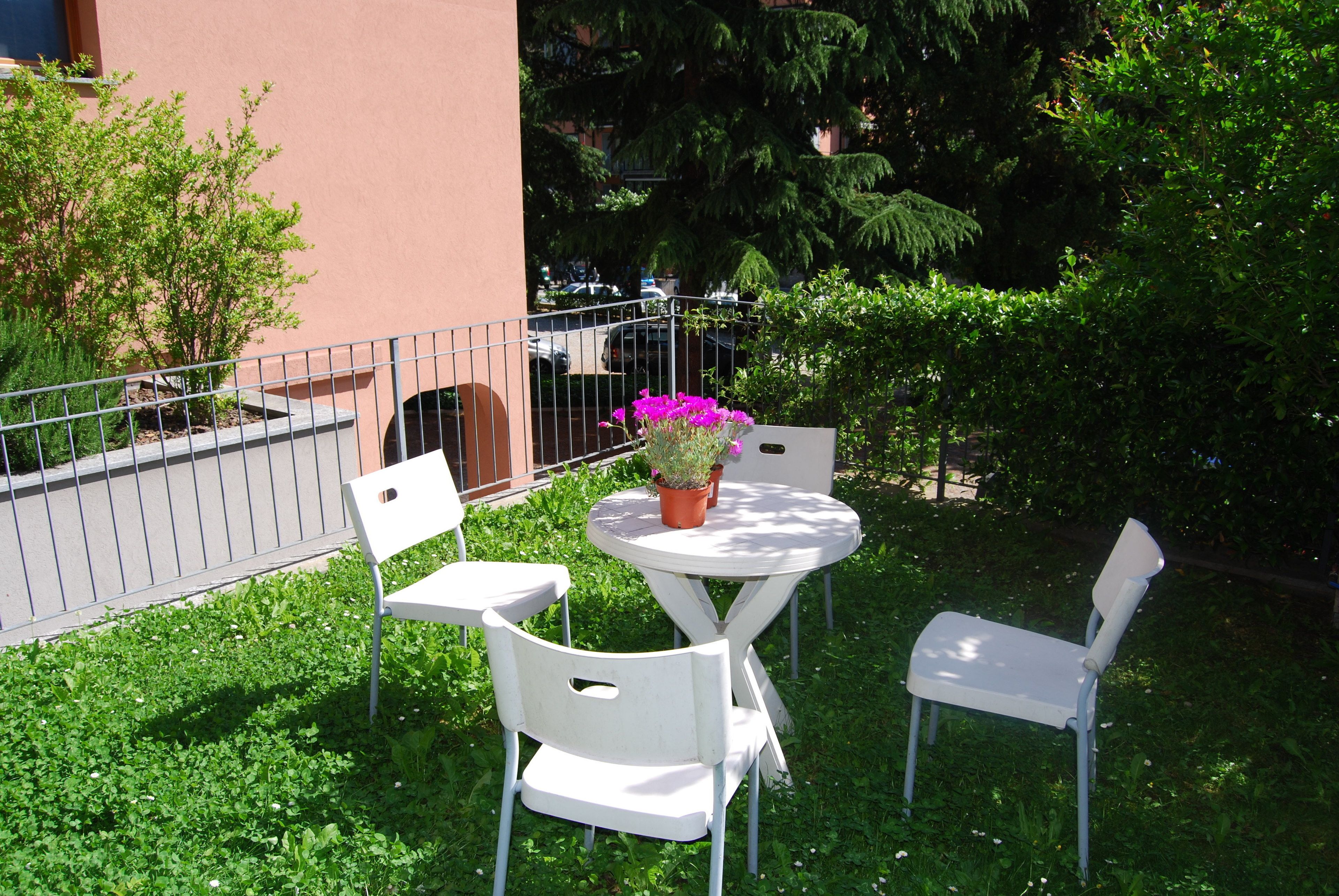 An outdoor seating area with a round table and two white chairs, surrounded by greenery and a metal fence.