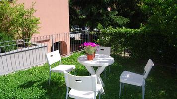 An outdoor seating area with a round table and two white chairs, surrounded by greenery and a metal fence.