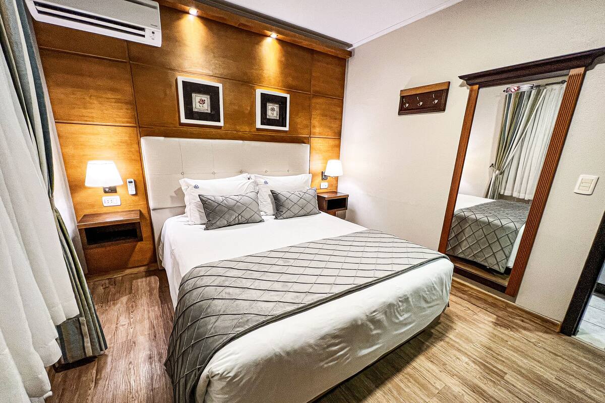A hotel room with a large bed, wooden headboard, two bedside lamps, a mirror, and a wall-mounted air conditioning unit.
