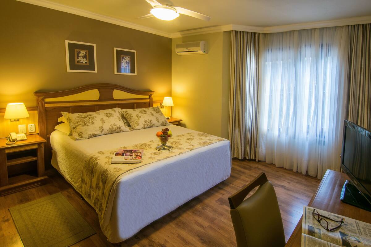 A hotel room with a bed, bedside lamps, a television, a desk with a chair, and a window with curtains.