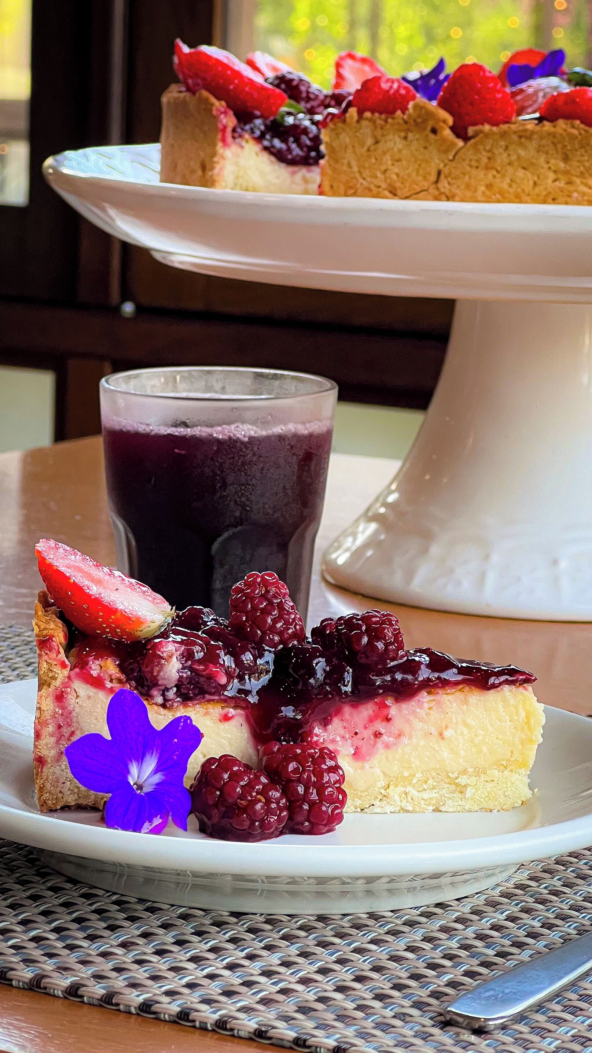 Ein Stück Käsekuchen mit Beeren auf einem Teller, ein Glas Beerengetränk und eine weiße Tortenplatte.