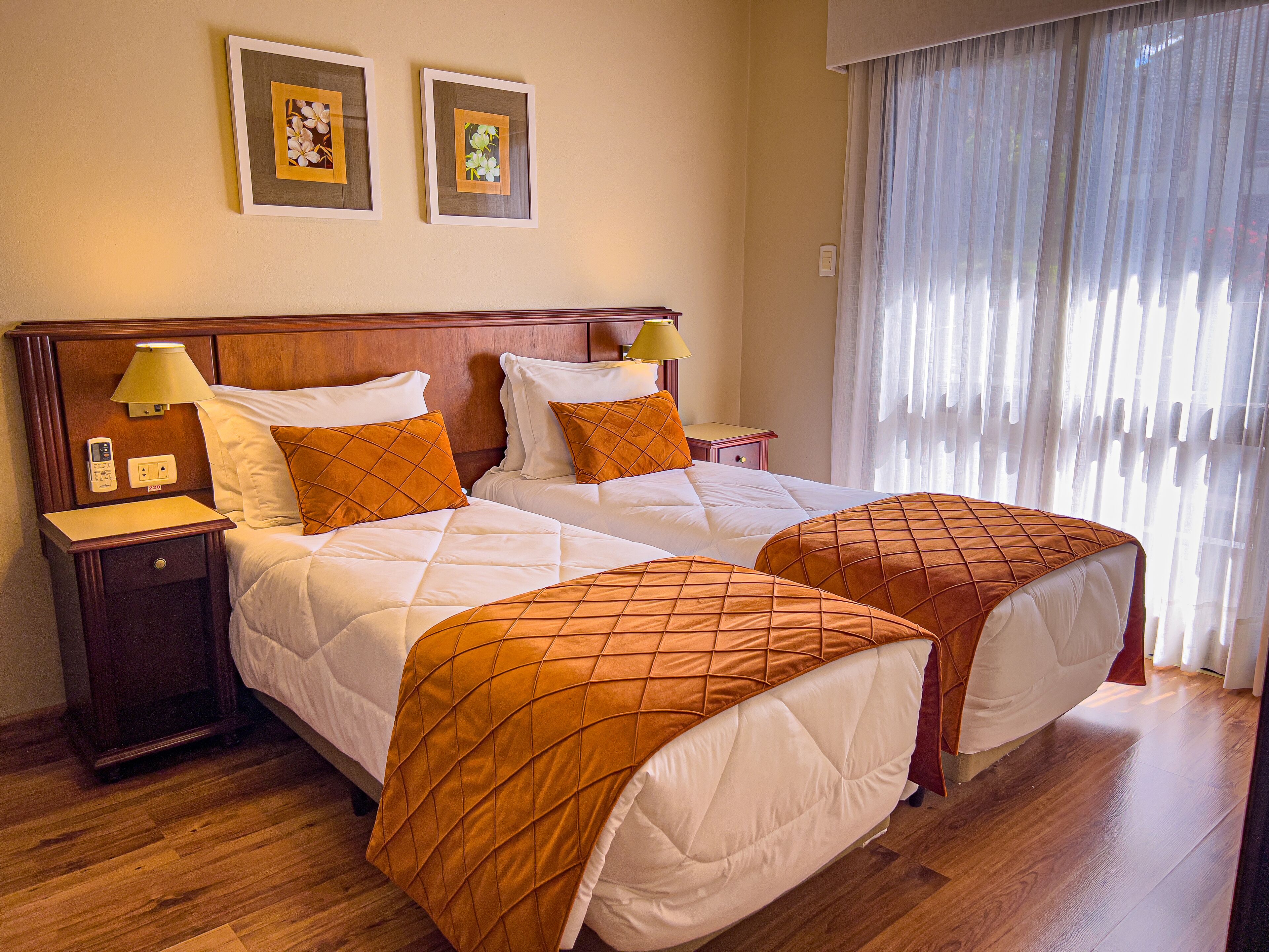A hotel room with two beds, wooden headboards, a nightstand with a lamp, and a window with sheer curtains.