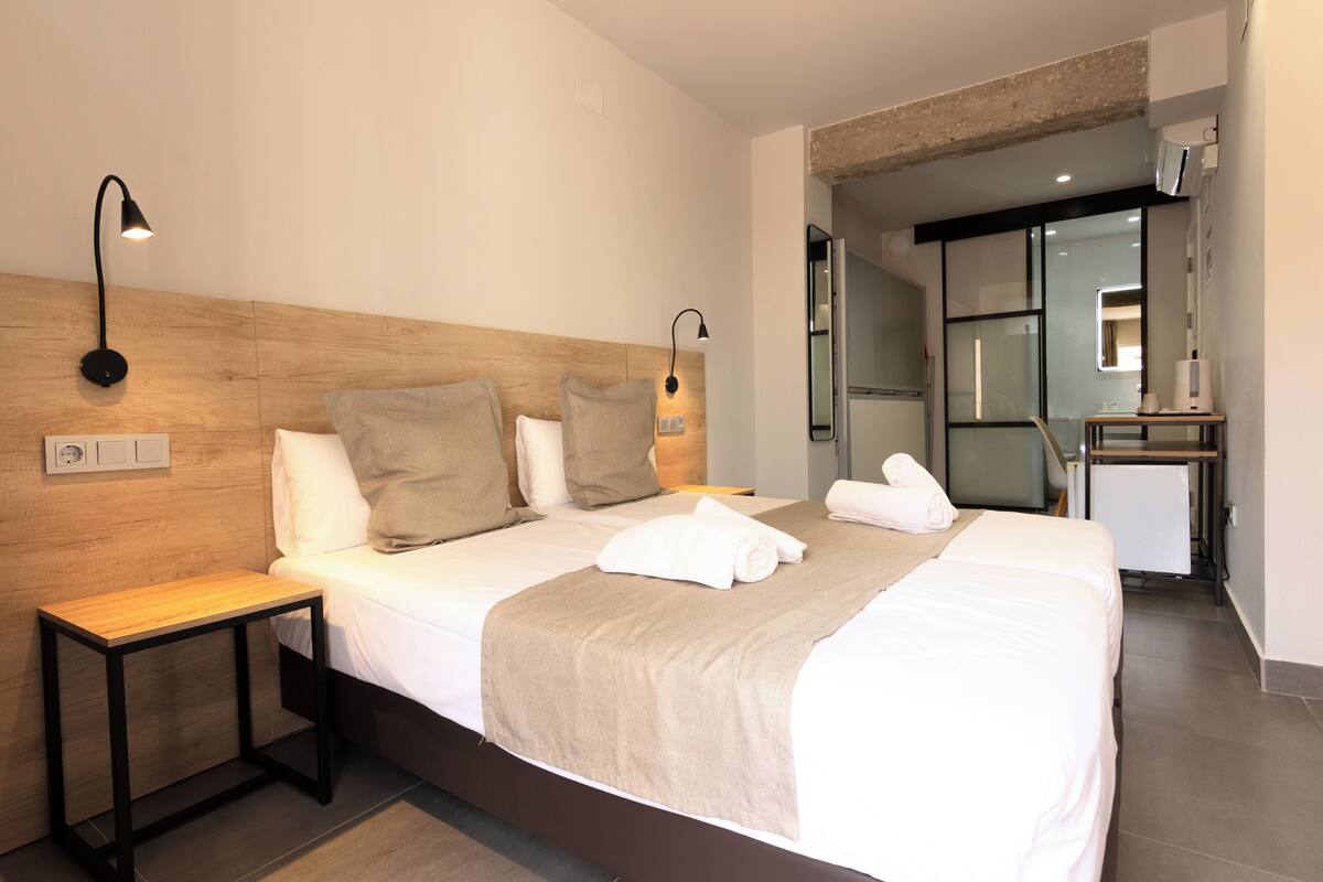 Moderni hotellihuone, jossa on puinen sängynpääty, yöpöytä, työpöytä tuolilla ja kylpyhuone, joka näkyy avoimesta ovesta.