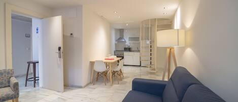 A modern living room with a blue sofa, a dining area with a table and chairs, and a kitchen in the background.