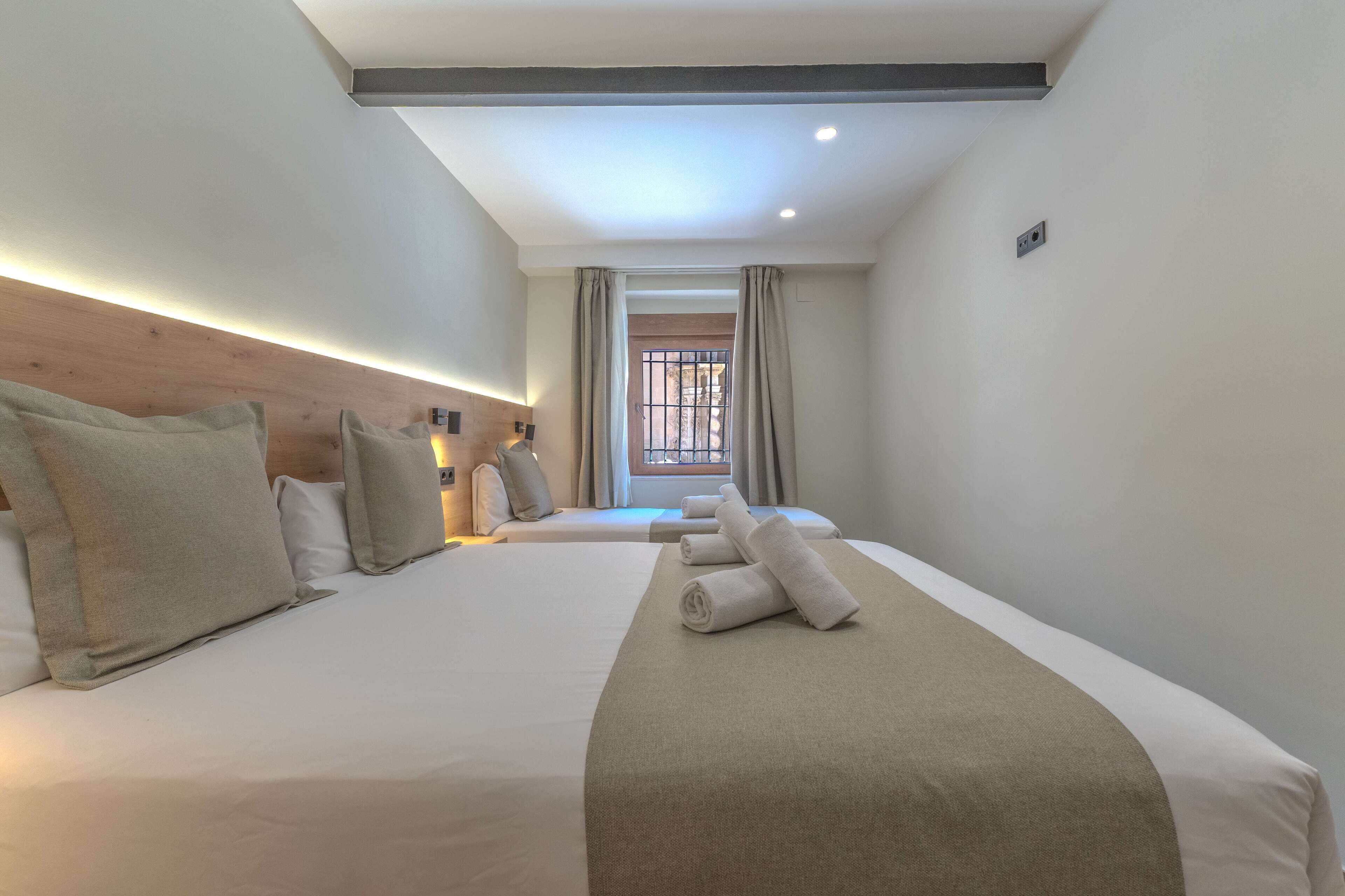Habitación de hotel con dos camas, cabecero de madera, ventana con cortinas e iluminación en el techo.