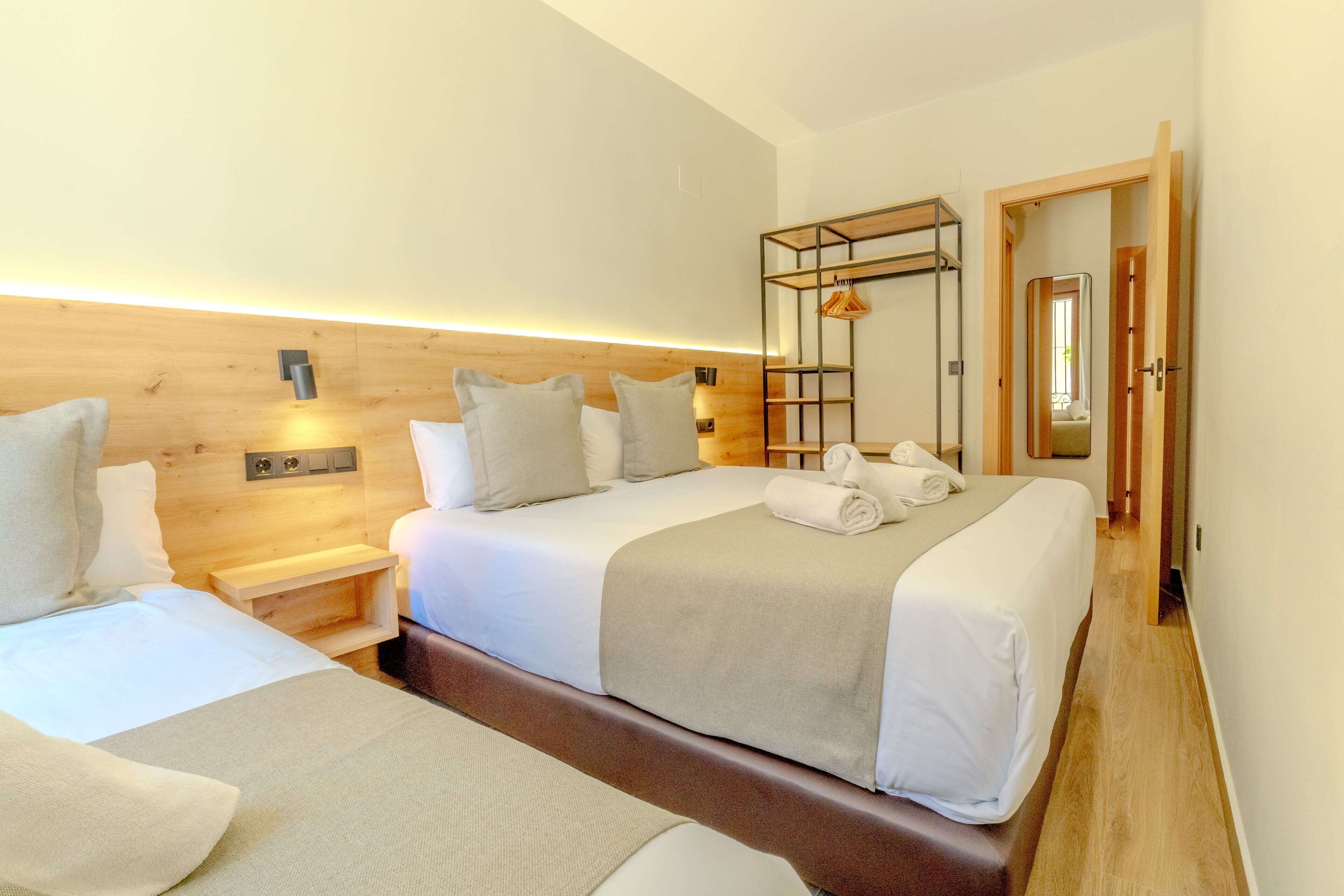 A hotel room with two beds, wooden headboards, a bedside table, a shelf, and a door leading to another room.