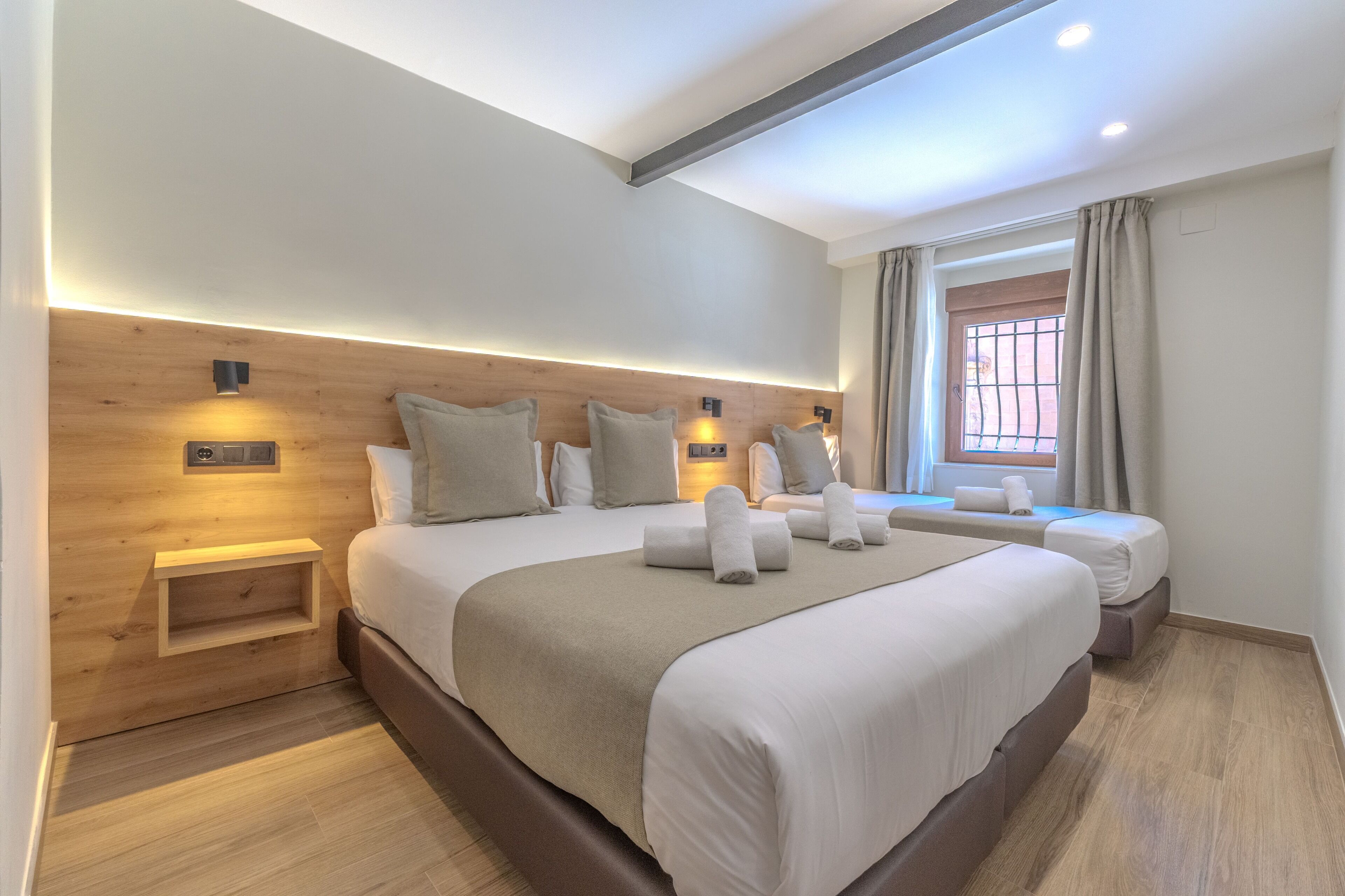 A modern hotel room with a large bed, bedside tables, a wooden headboard, and a window with curtains.