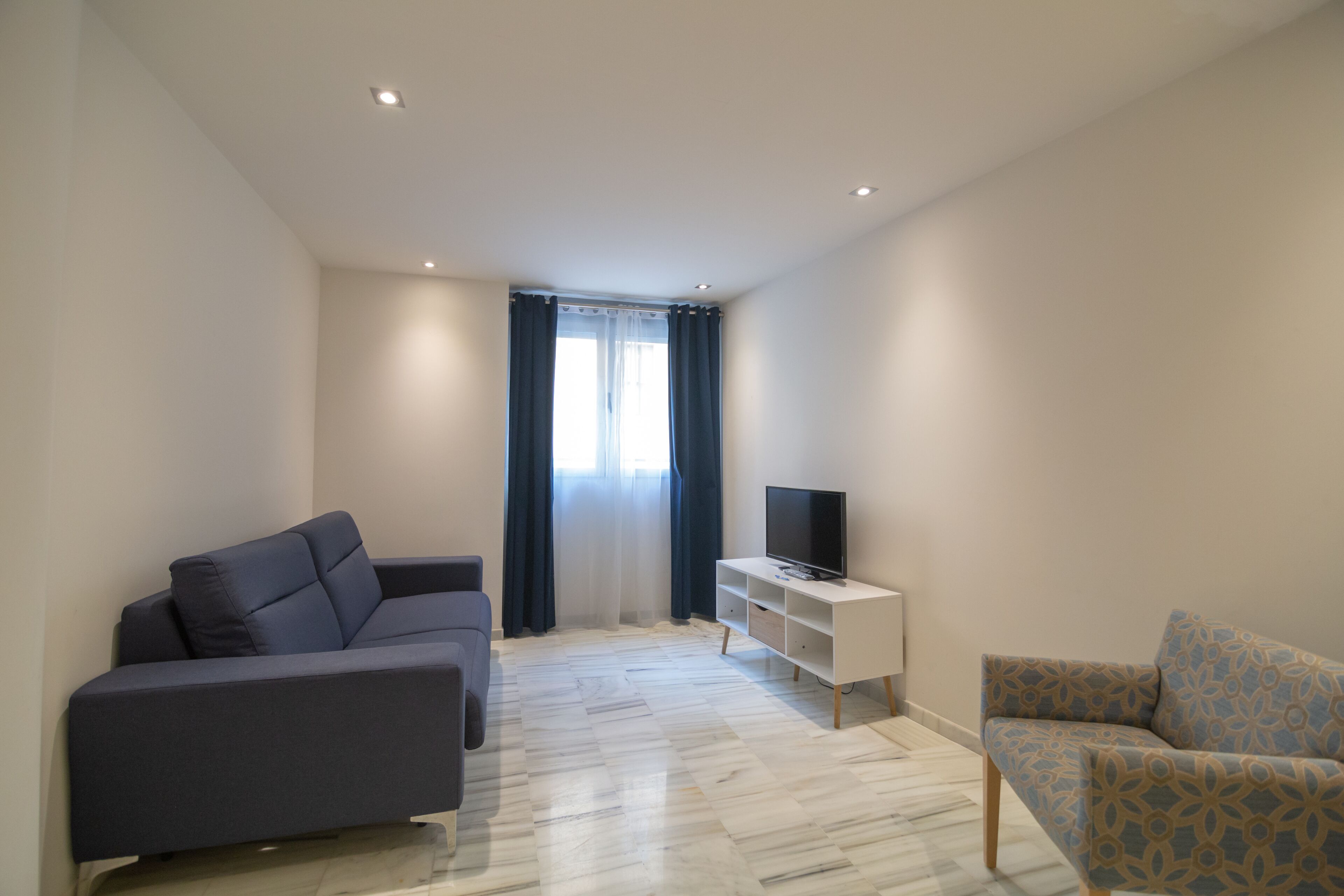 A modern living room with a grey sofa, a patterned armchair, a TV on a white stand, and a window with curtains.