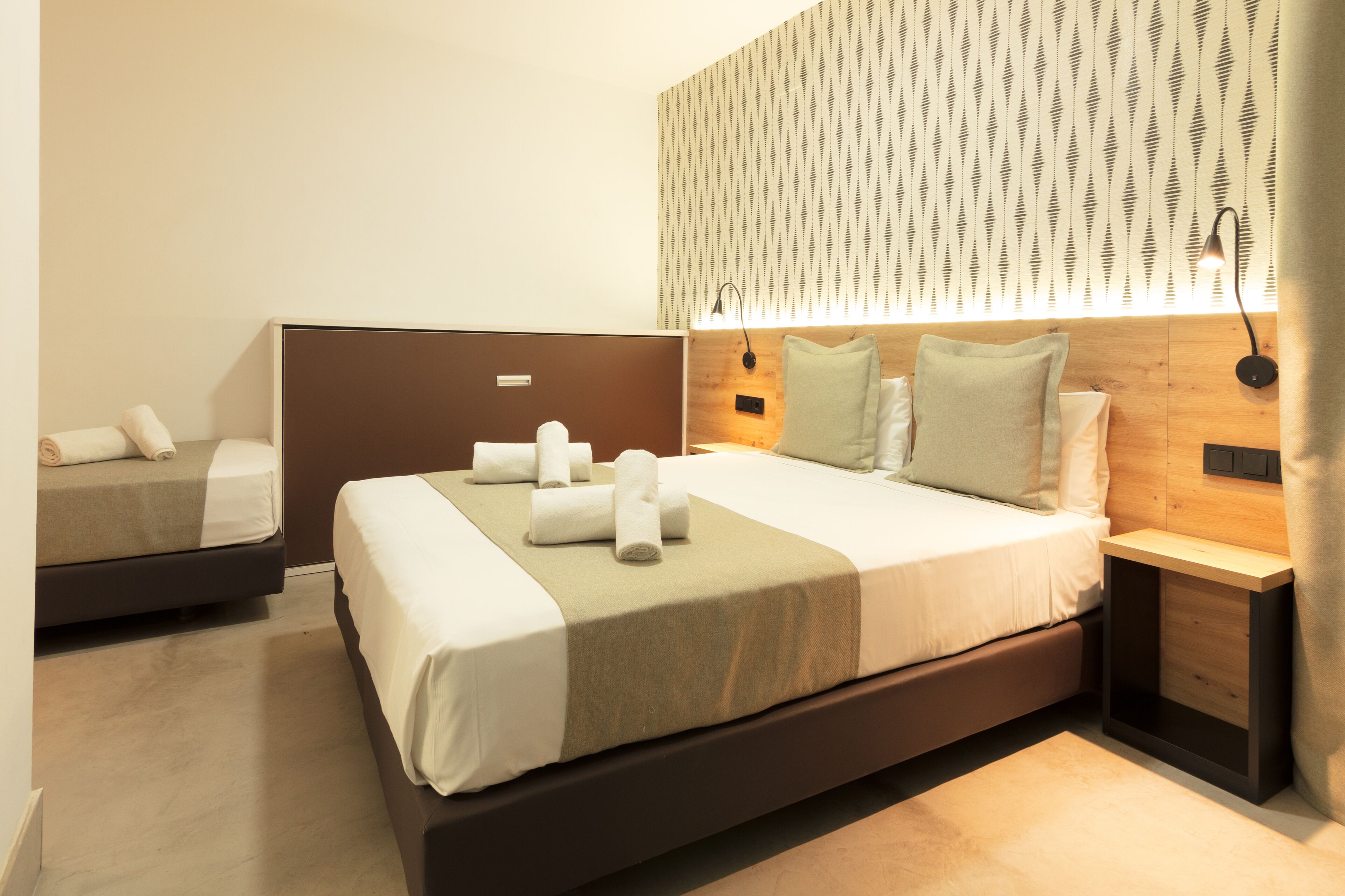 A hotel room with a large bed, a smaller bed, a nightstand, a lamp, and a wall-mounted light fixture.