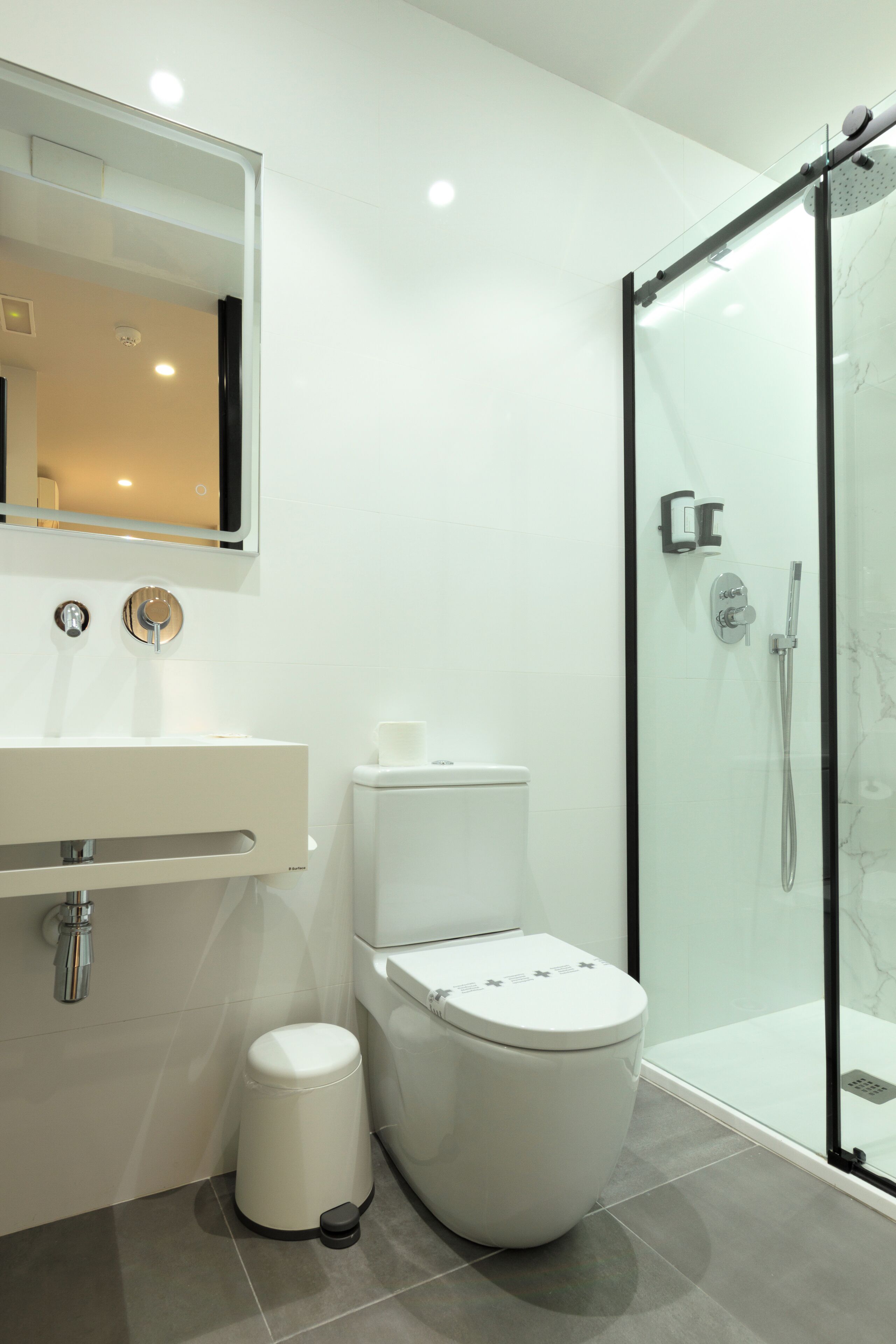 Une salle de bain moderne avec une douche à parois de verre, des toilettes blanches et un lavabo surmonté d’un miroir.