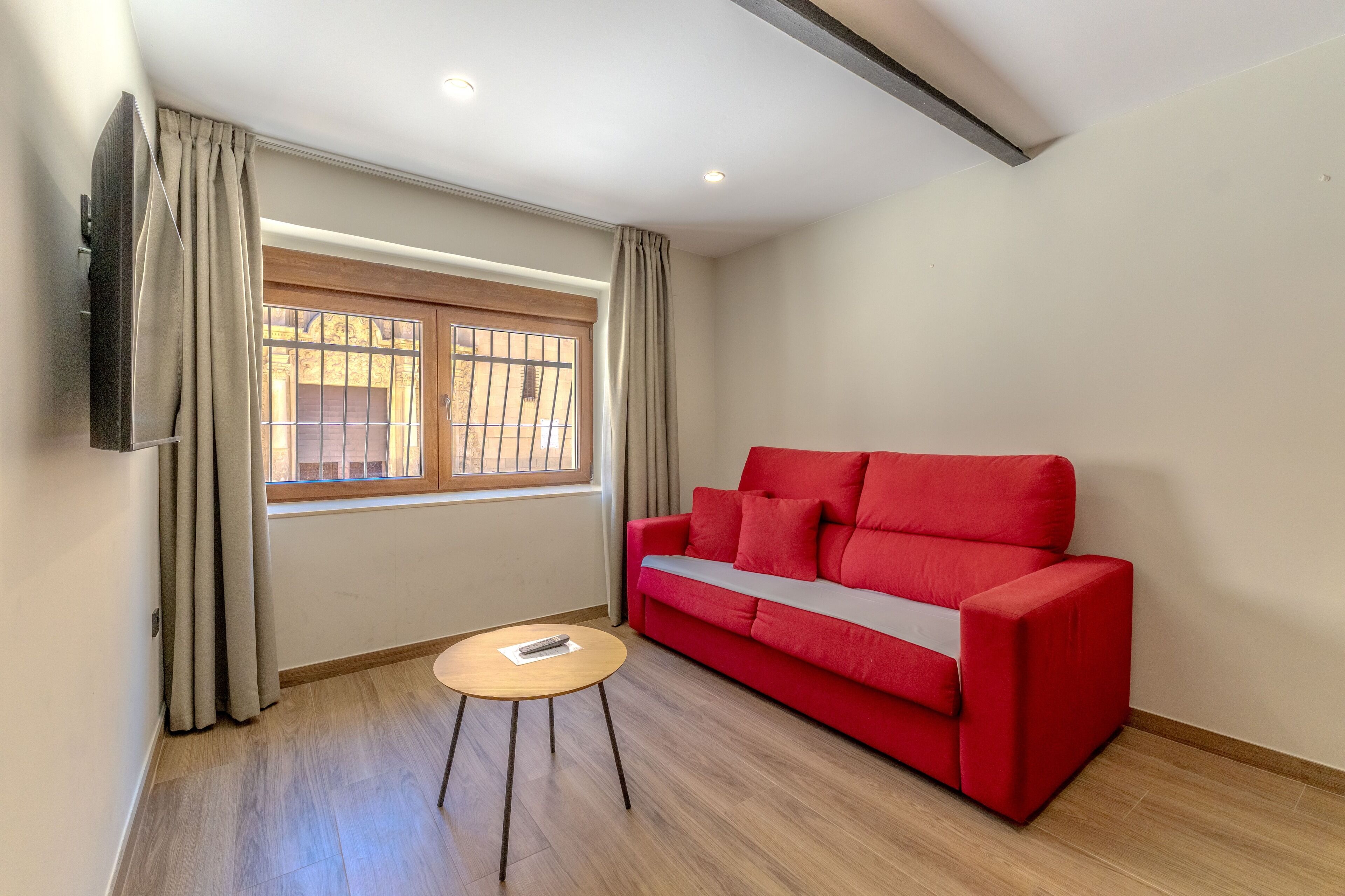 Una habitación con un sofá rojo, una pequeña mesa redonda, un televisor montado en la pared y una ventana con cortinas.