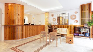 A reception area with a wooden reception desk, a sofa, and a coffee table. There are chairs and a small wooden box on the floor. - Hotel La Milagrosa (Alicante)