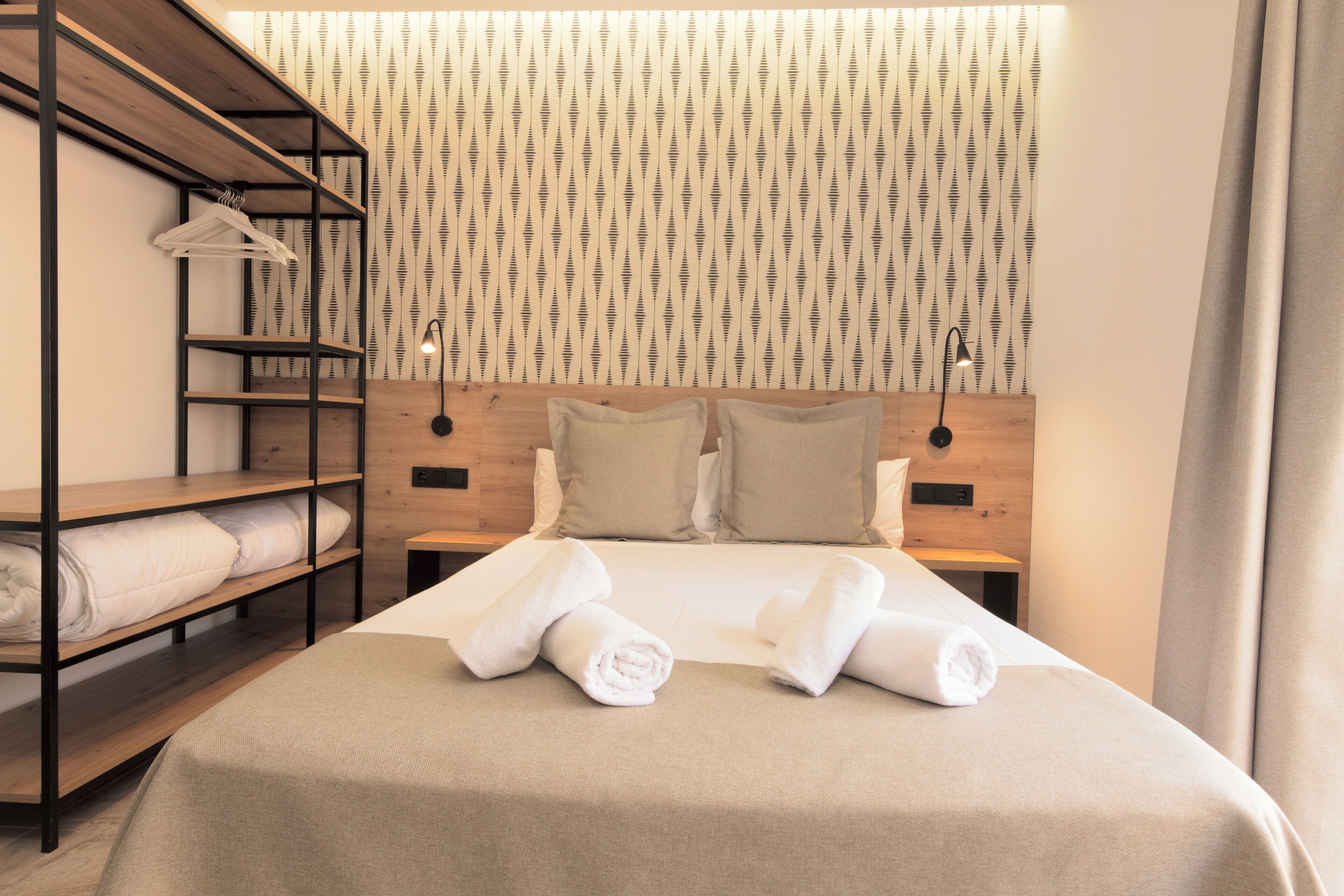 A neatly made bed with pillows and rolled towels, a wooden shelf unit with folded towels, and a wall with a striped pattern.