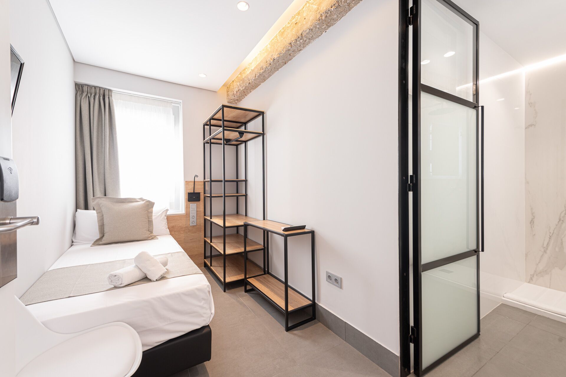 A modern hotel room with a bed, a built-in bench, a glass door, and a wooden ceiling.
