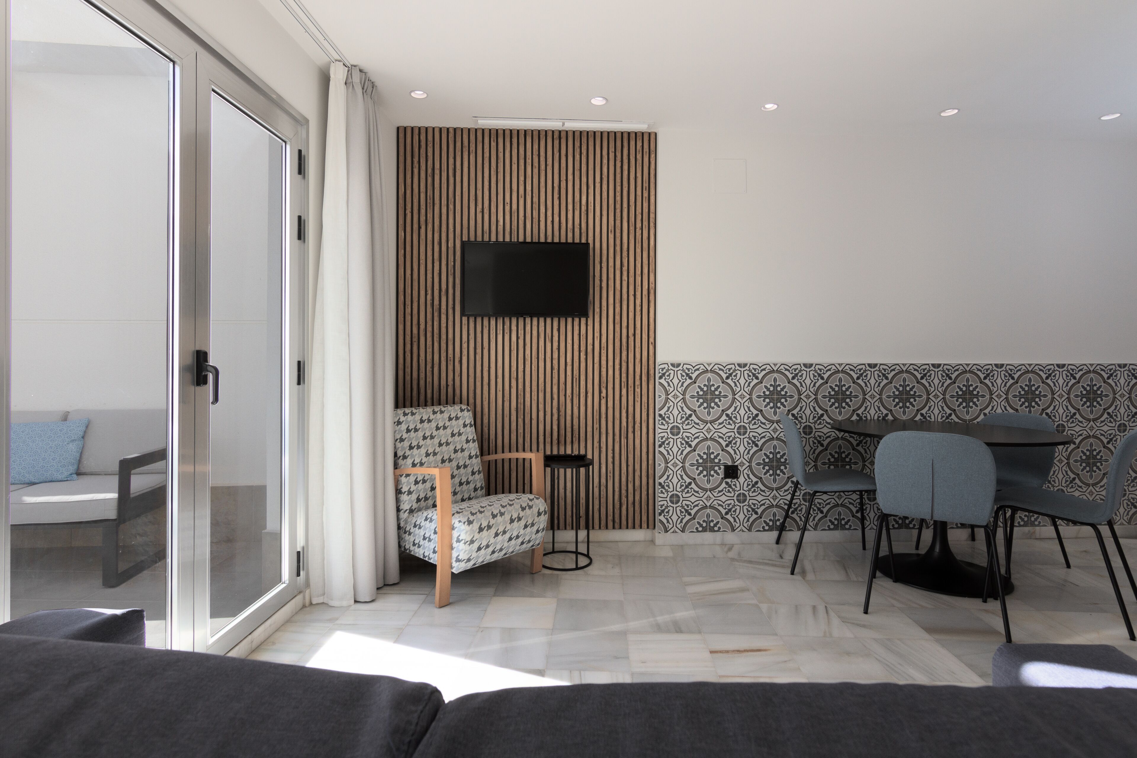 Una sala de estar moderna con un ventanal que da a un balcón, una zona de estar con un sofá y una silla, un televisor de pantalla plana montado en una pared de paneles de madera, y un comedor con mesa y sillas.