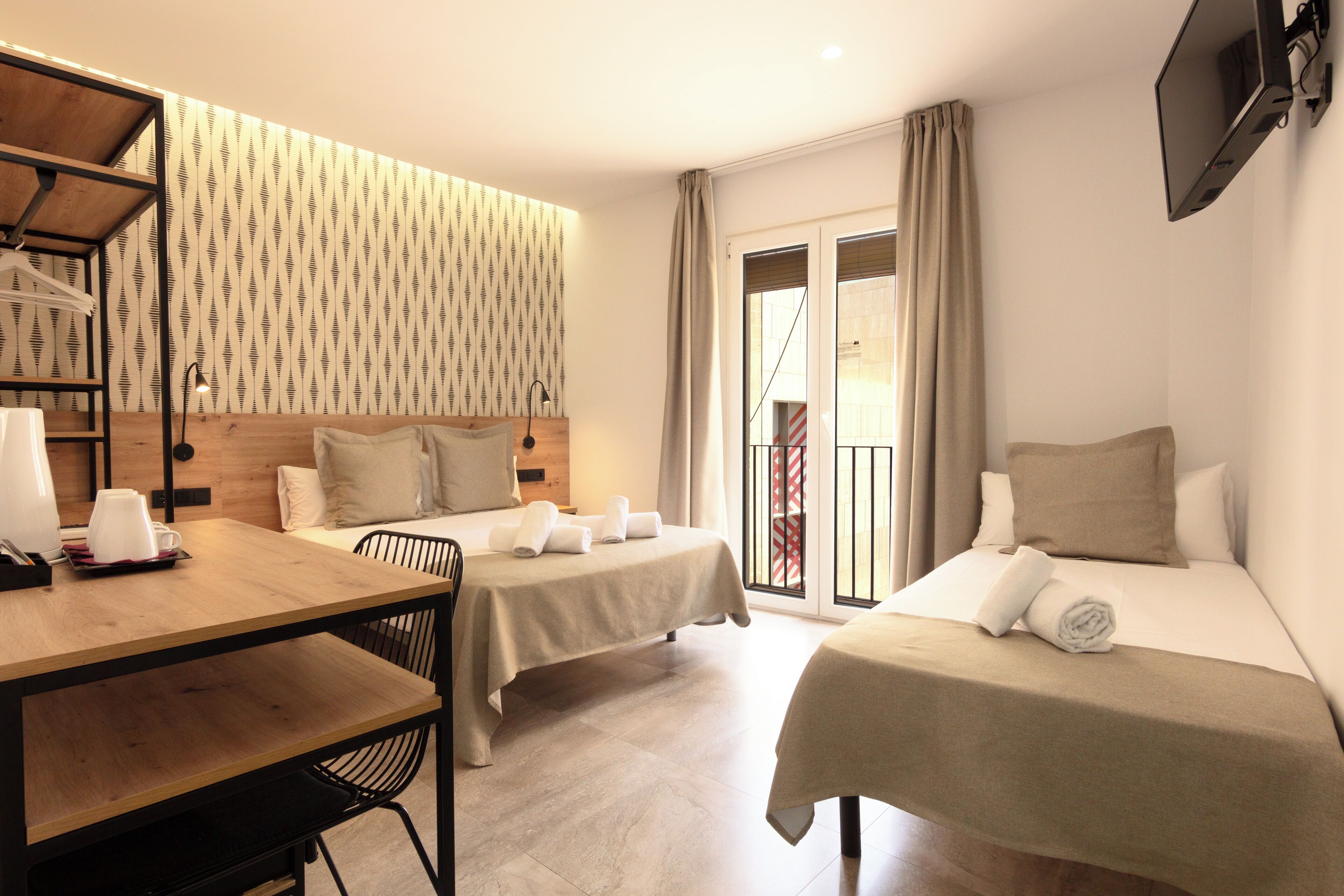 A hotel room with two beds, a desk, a chair, a TV, and a patterned wall.