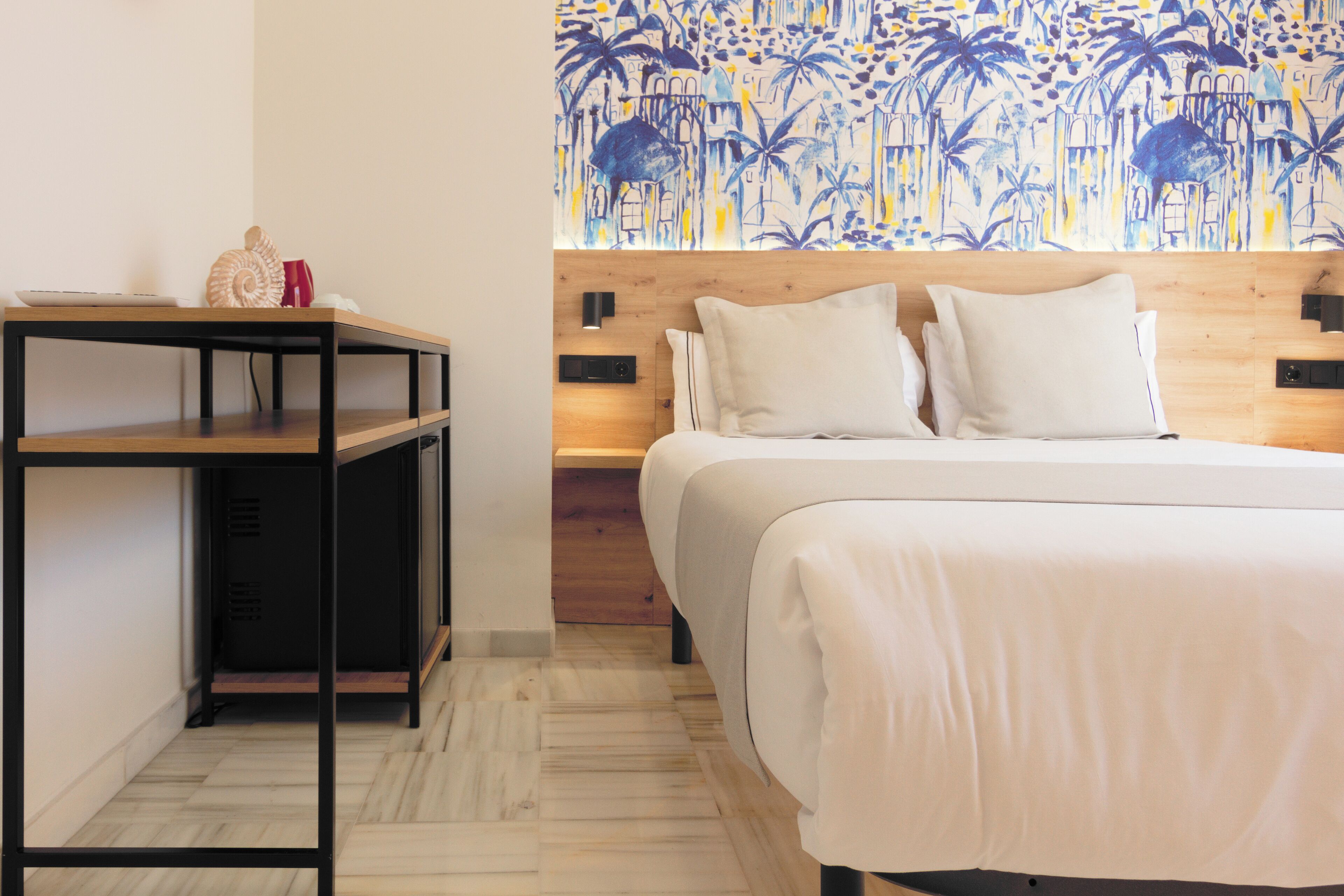 A hotel room with a wooden headboard, a bed with white bedding, a small table with a decorative item, and a wall with a blue and yellow abstract pattern.