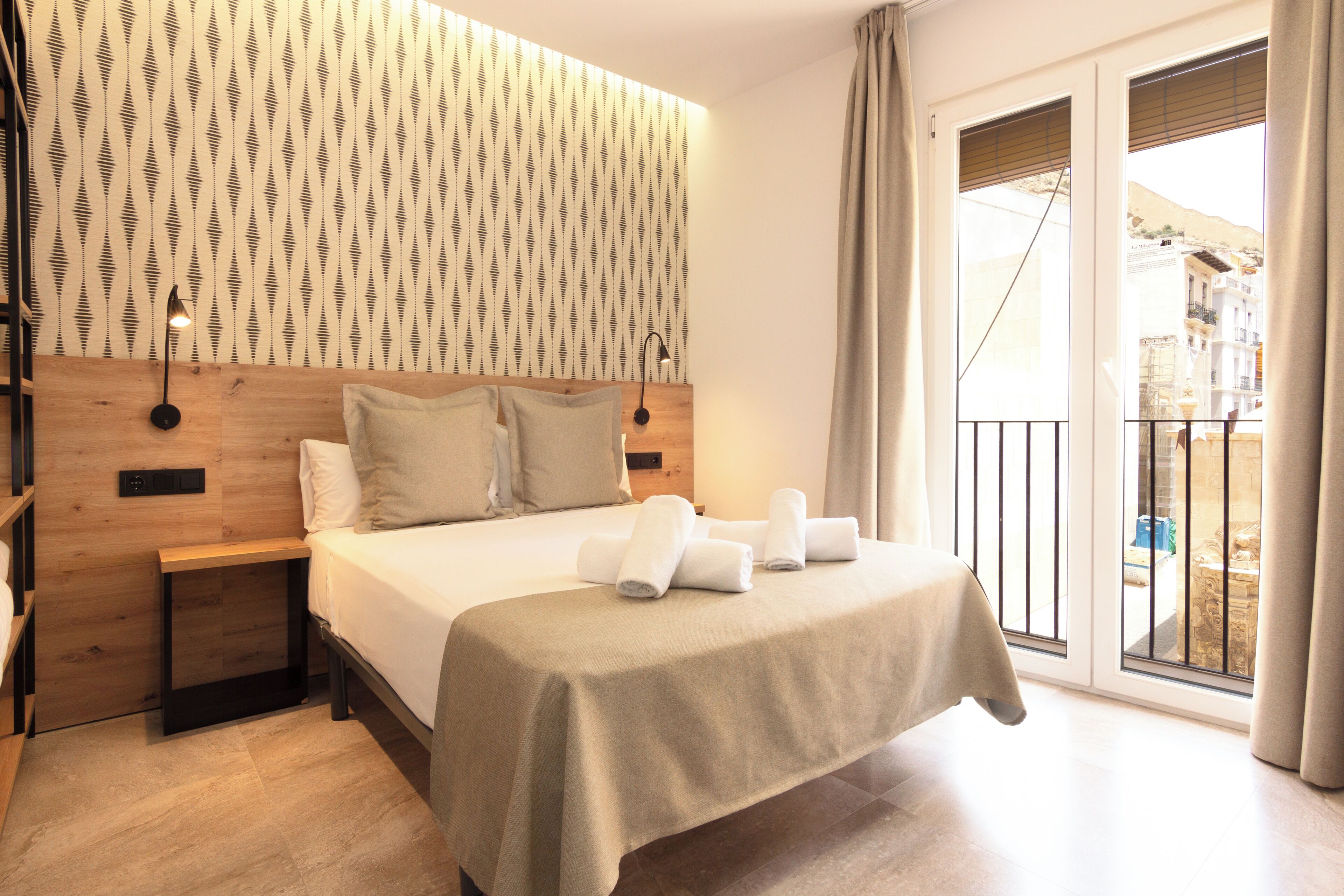 A hotel room with a bed, a nightstand, a wall-mounted lamp, and a view of the outside.