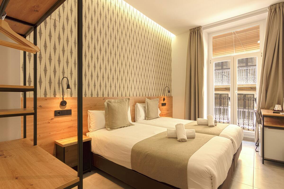 Hotellihuone, jossa on kaksi sänkyä, puinen pääty, yöpöytä, työpöytä ja ikkuna, josta on näkymä parvekkeelle.