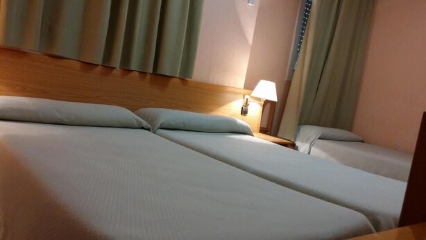 A hotel room with two beds, a wooden headboard, a nightstand with a lamp, and curtains. - Hotel Rialto (Alicante)