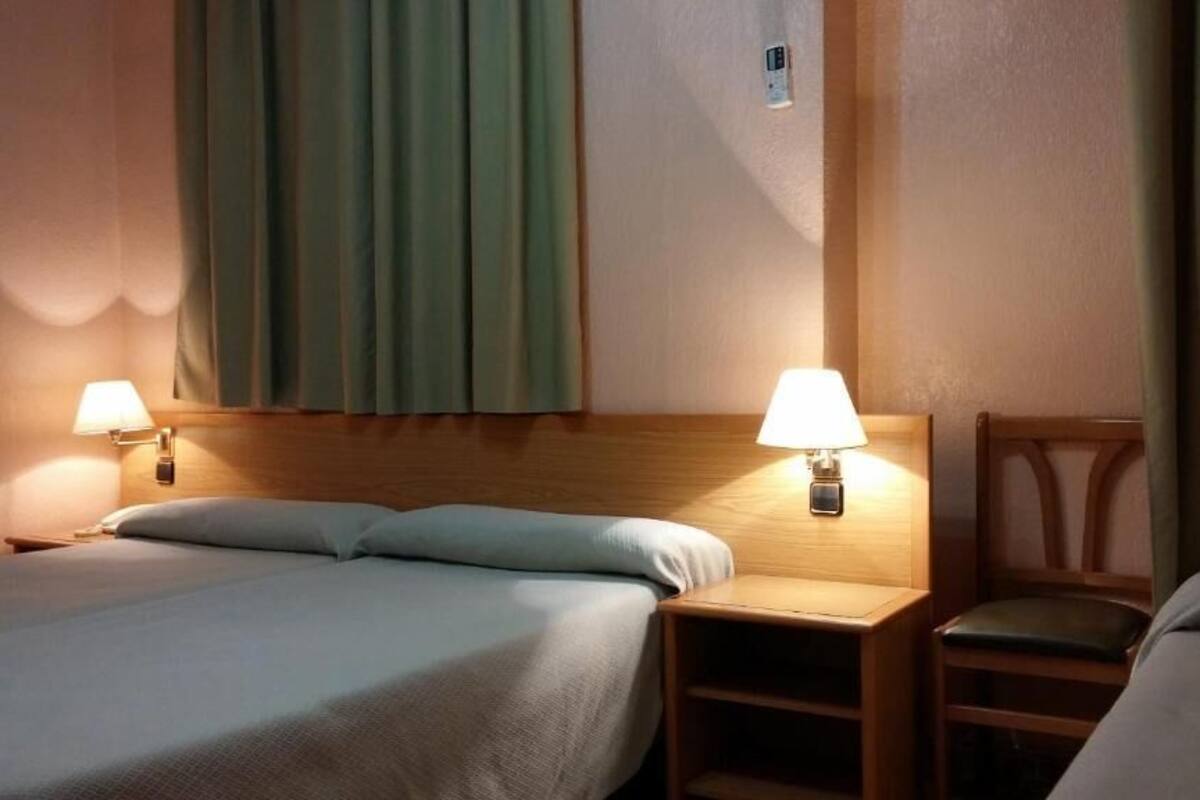 A hotel room with two beds, a nightstand with a lamp, a chair, and a window with curtains.